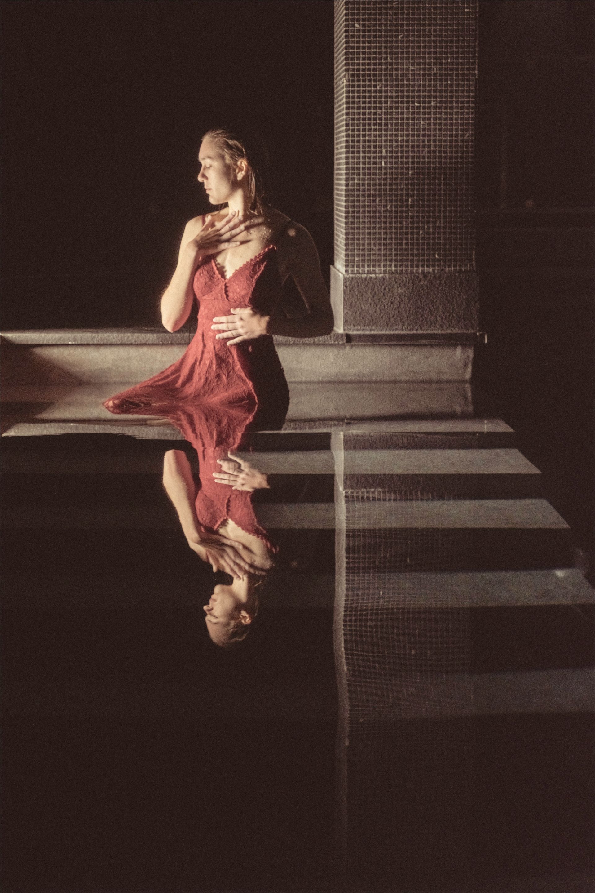 Woman in red dress by water, reflected in water; evening light.