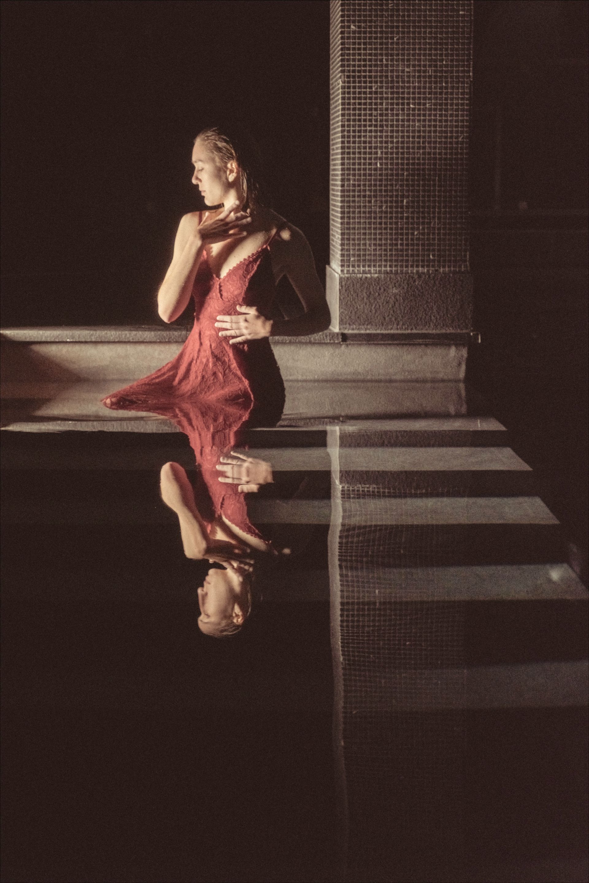 Woman in red dress, reflected in water, stands near brick wall.