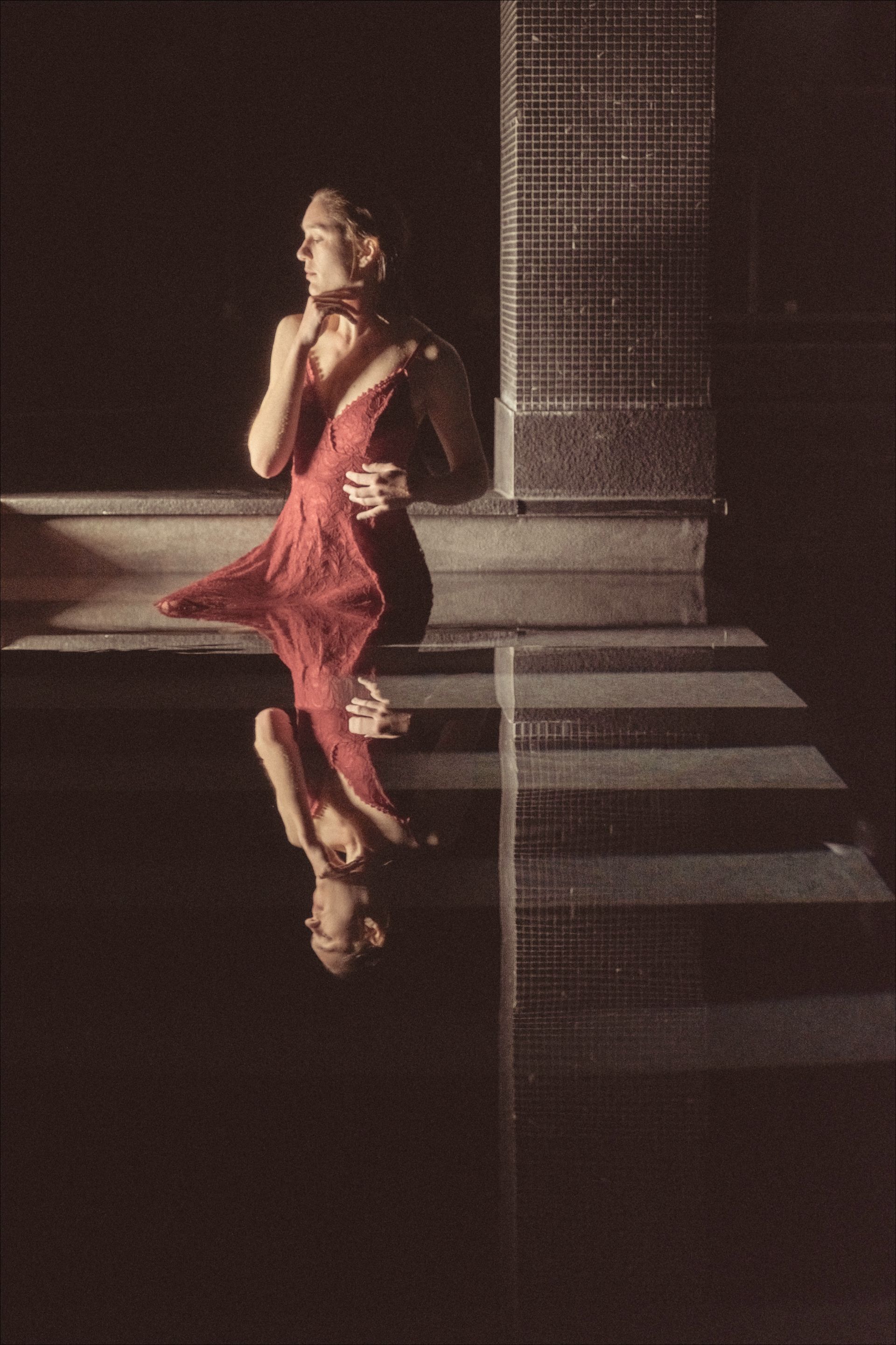 Woman in red dress reflected in water, standing by a tiled wall and steps.