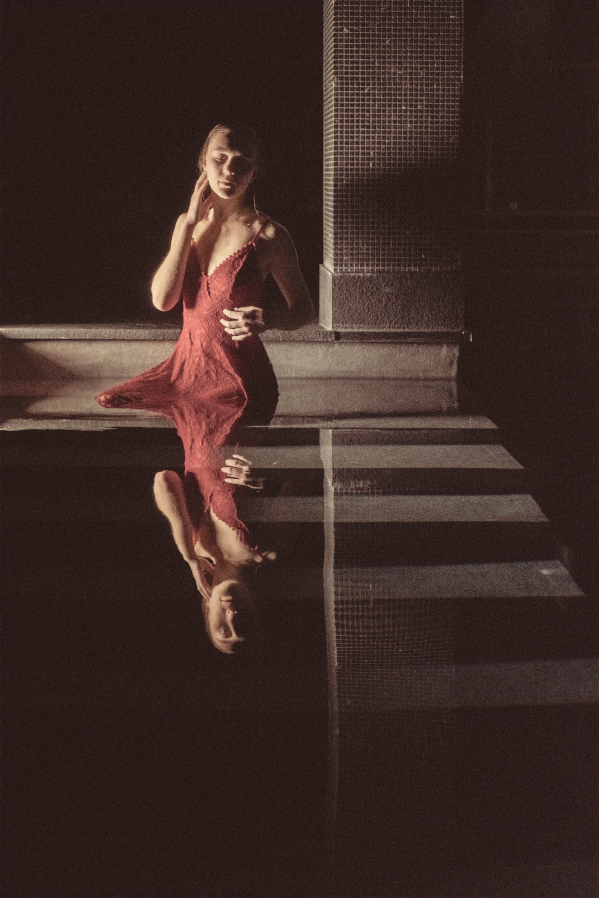 Woman in red dress sits by water, her reflection visible. Dark background.