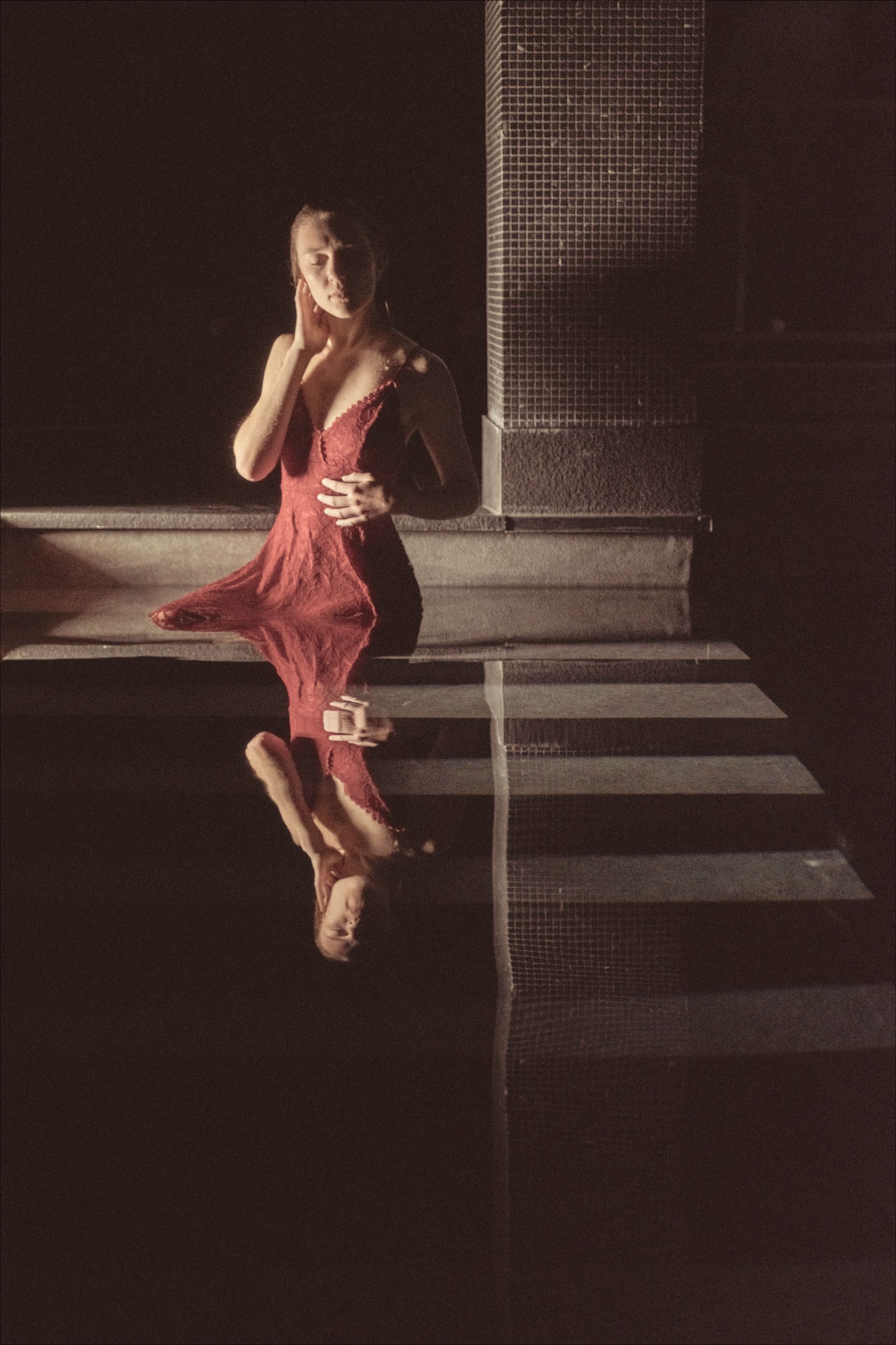 Woman in red dress sits near steps, reflection in water, brick wall in background.