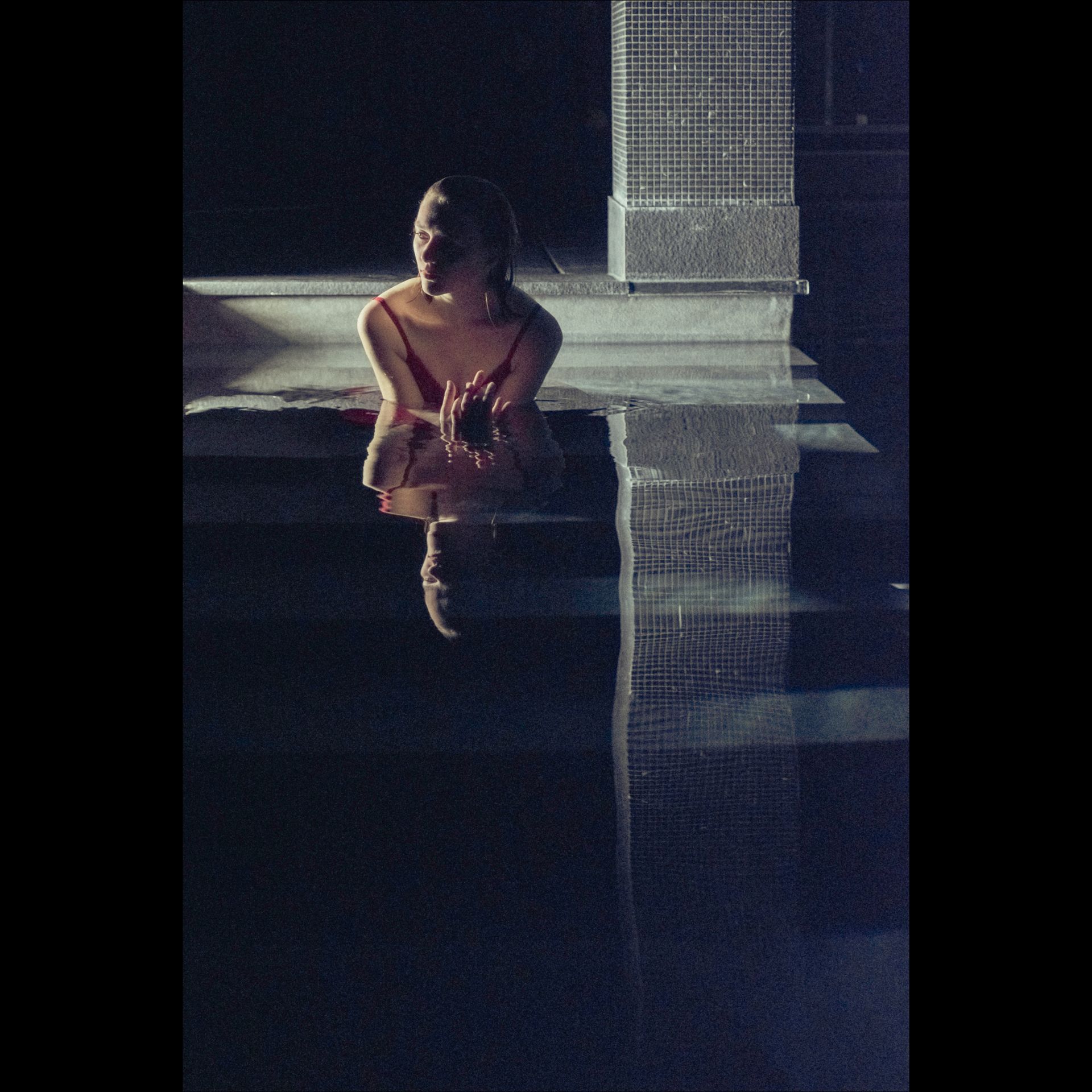 Woman sitting on edge of pool at night, reflection in water, dark setting.