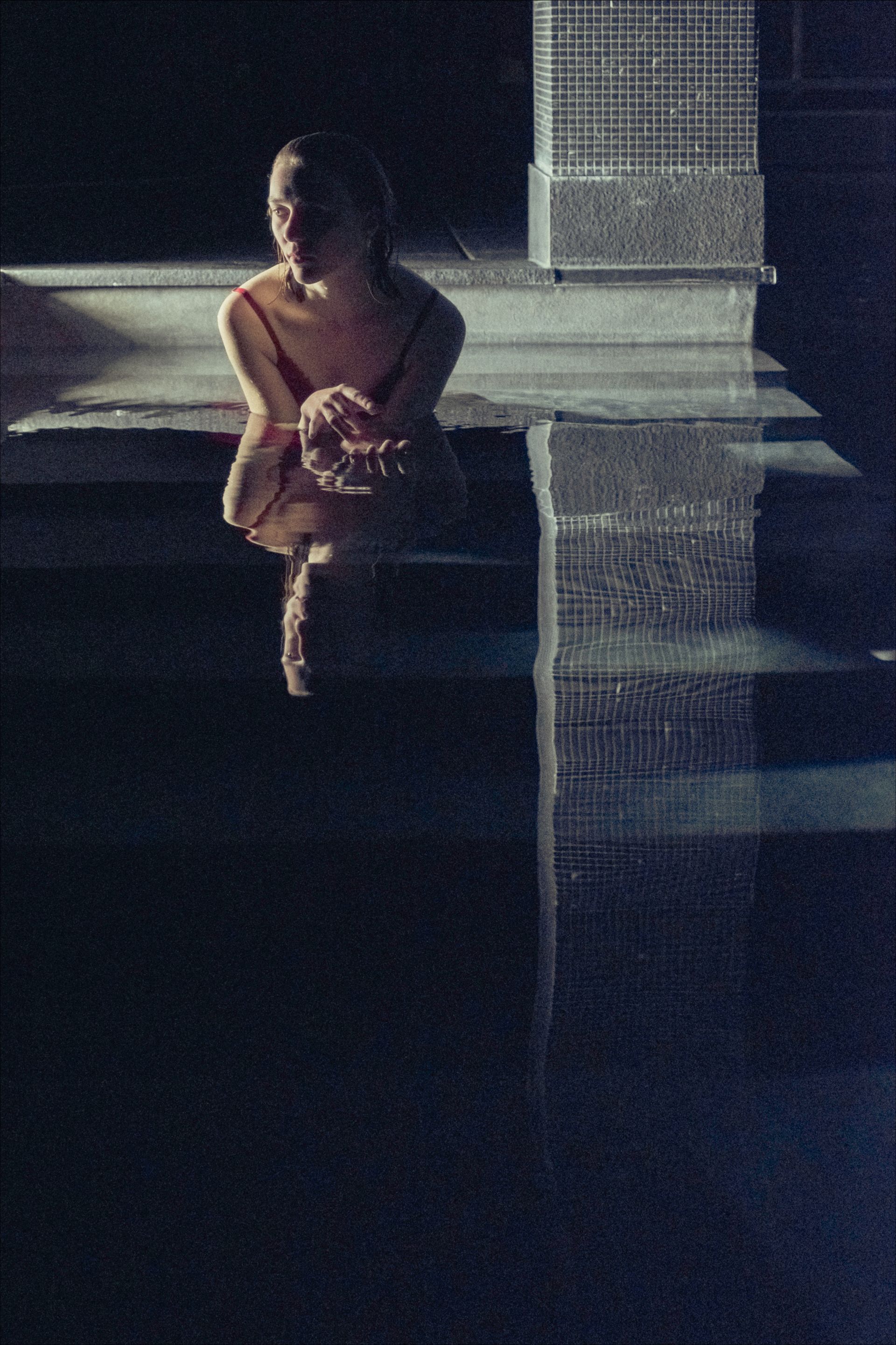 Woman in red swimsuit sits at edge of dark pool, reflecting in the water, under dim light.