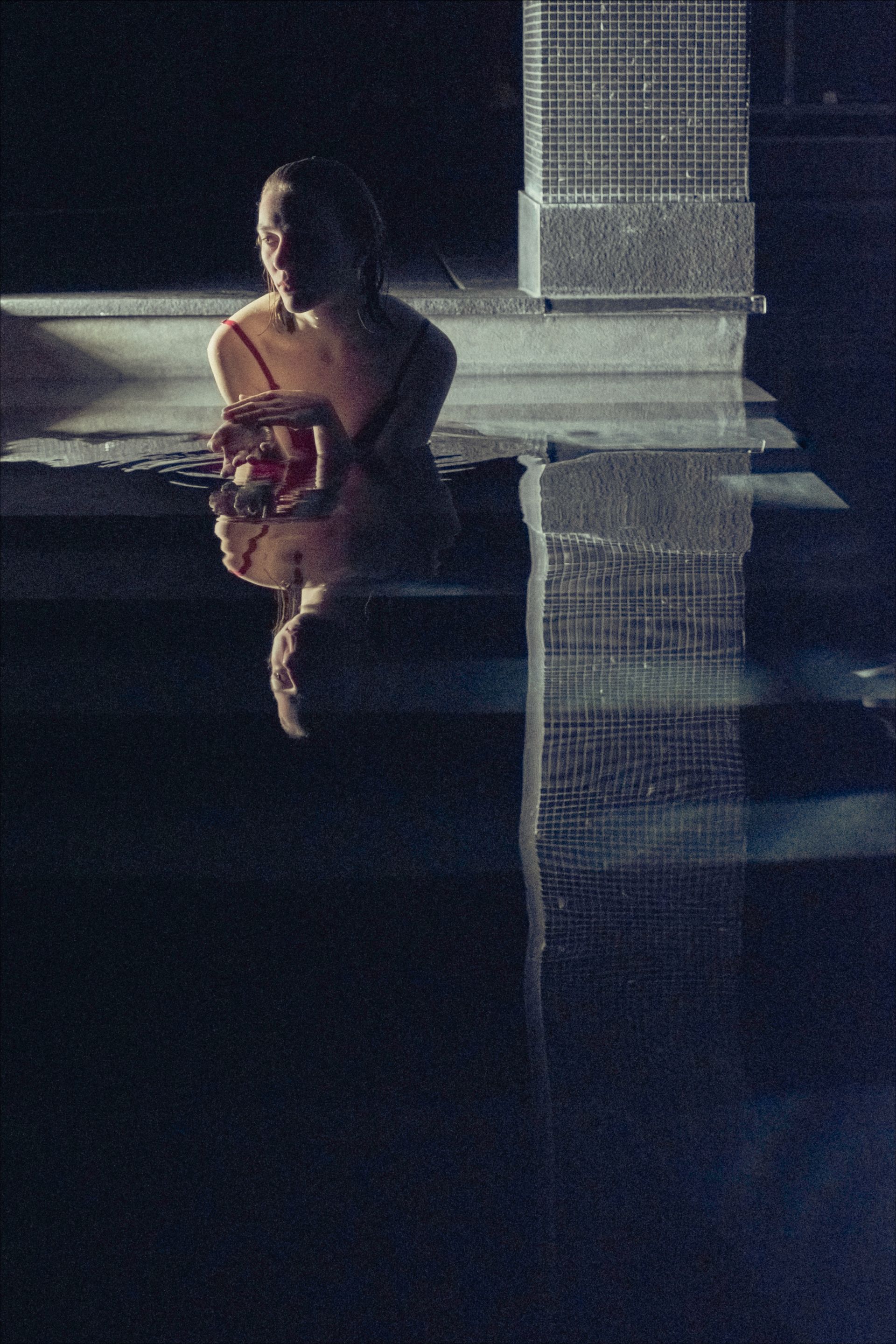 Woman in red swimsuit sits at edge of dark pool, reflecting in the water.