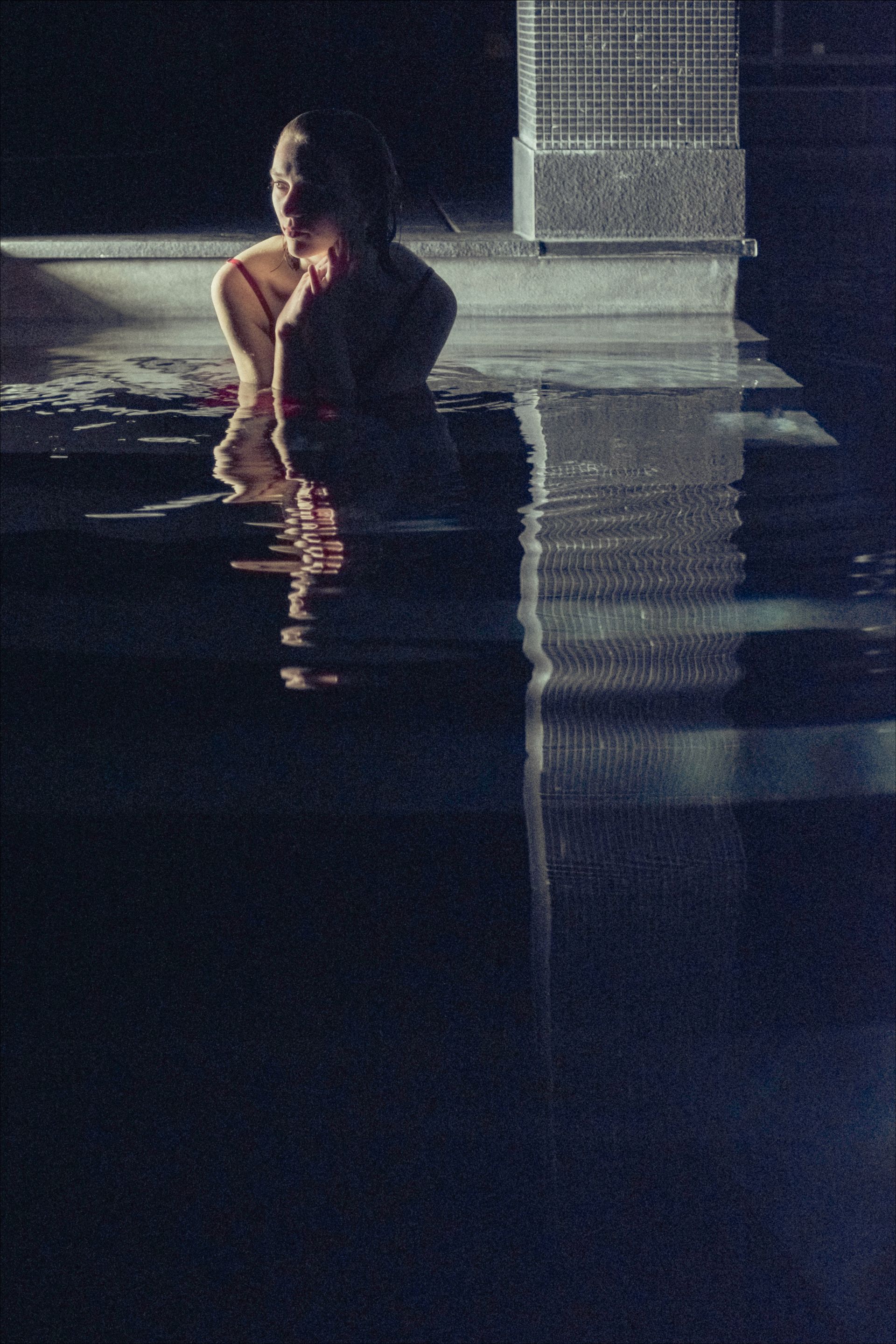 Person in shallow water, hands near face. Dark, possibly indoor setting; water reflects light.