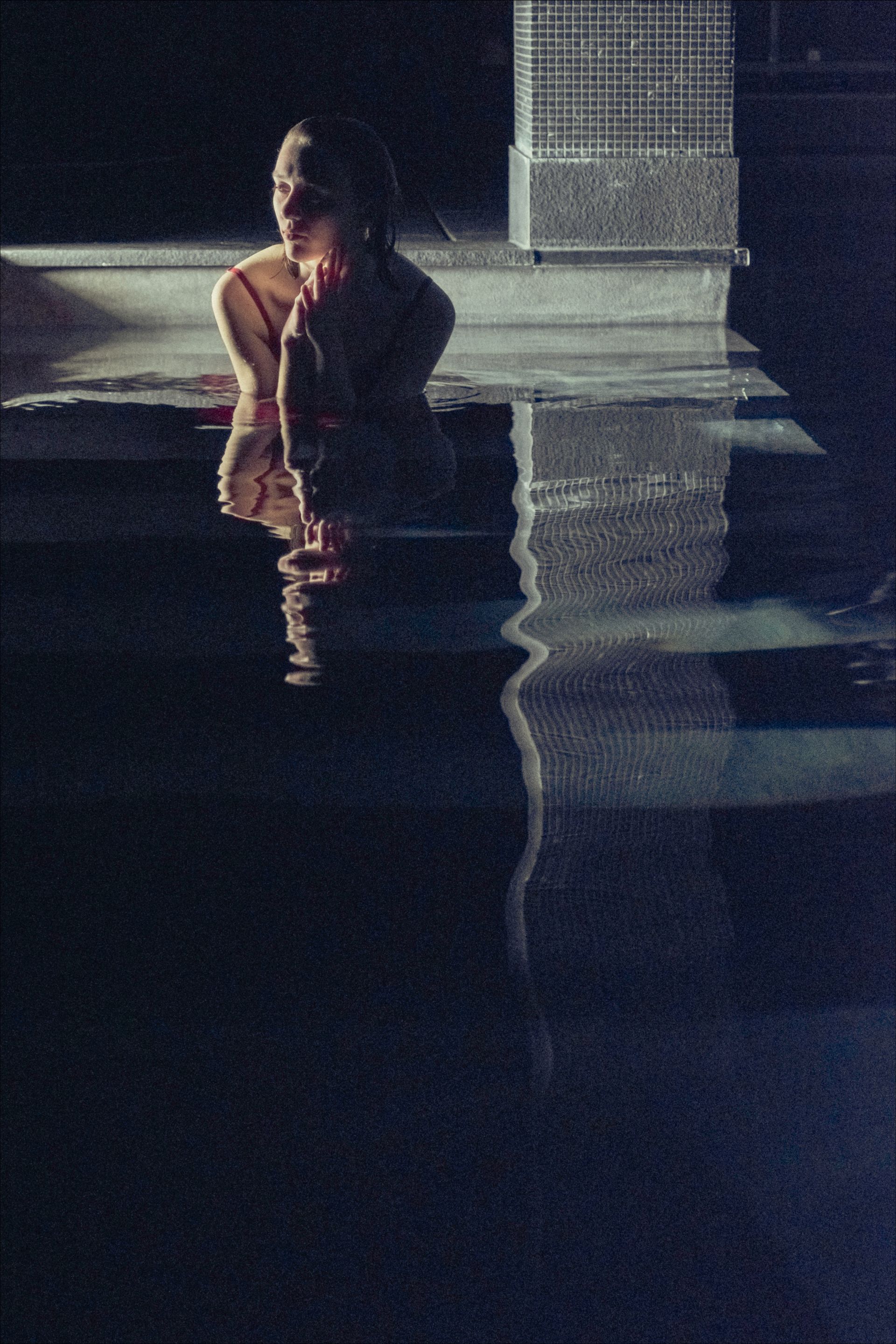 Woman in a pool, reflected in the water, looking upward. Dark setting, blurred and indistinct.