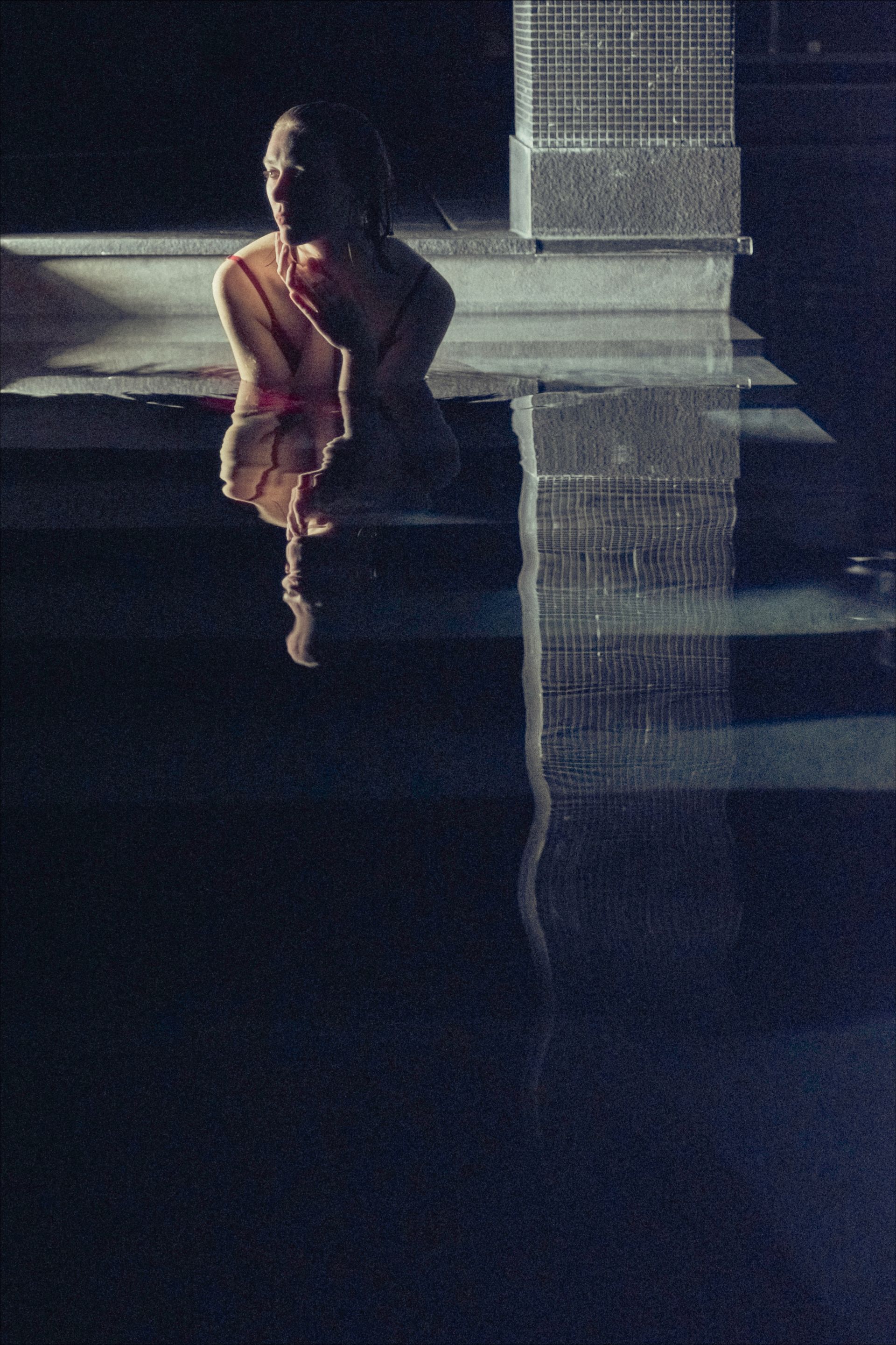 A person sits near water, reflecting in its surface. A building is also reflected. Dark and pale tones.