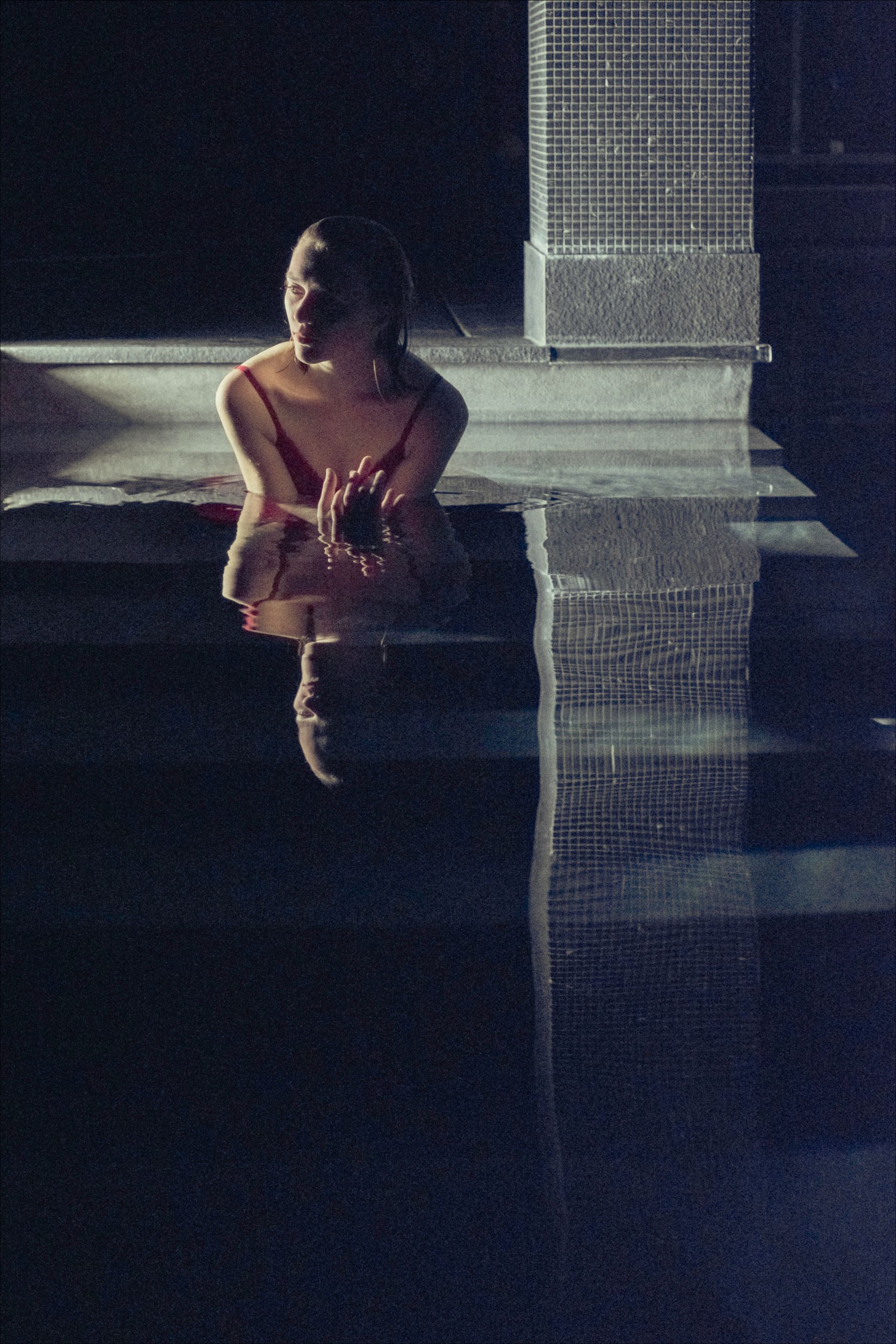 Woman in red dress sits by pool, gazing down. Dark blue water and night scene.
