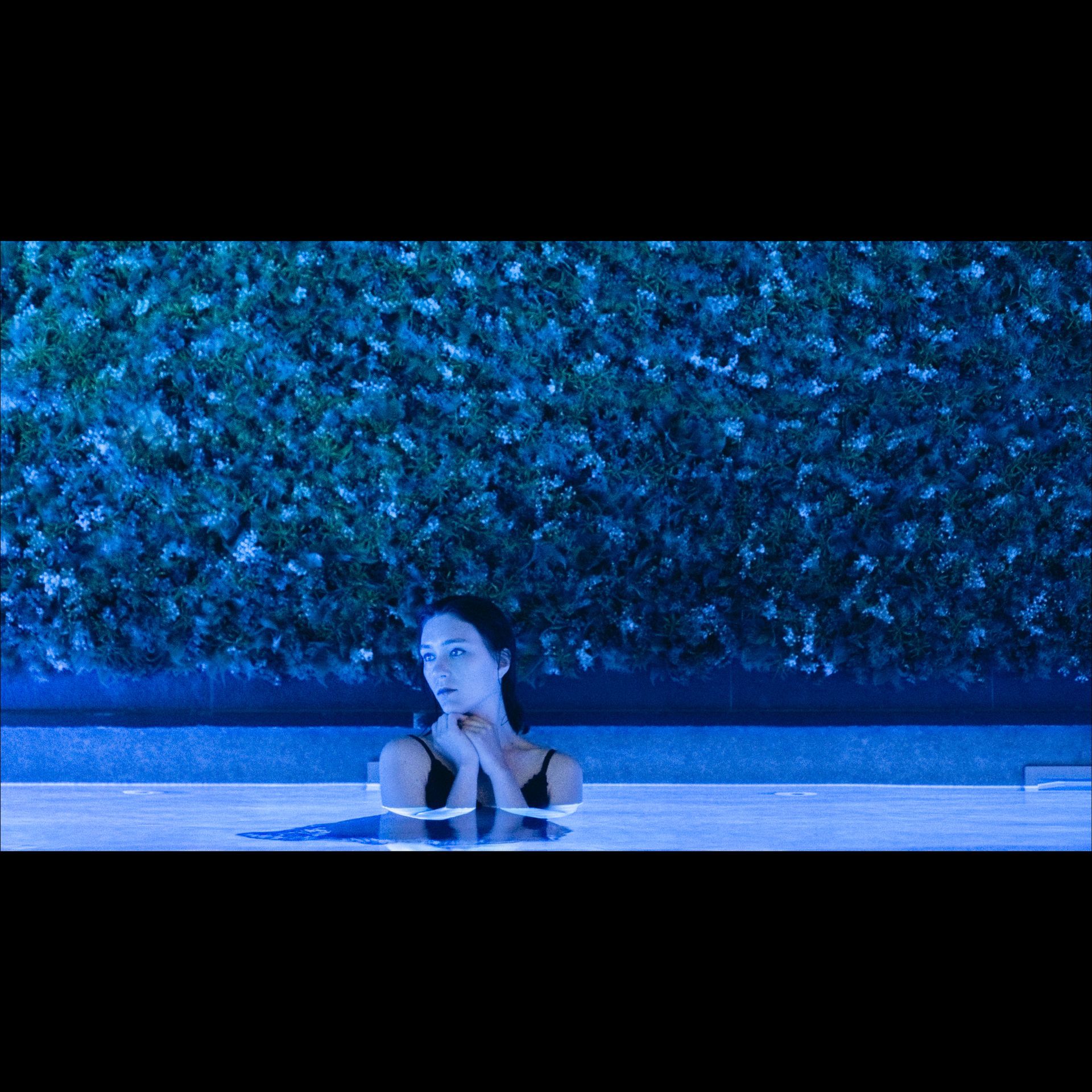 Woman in a pool, surrounded by blue light. Dark hair, looking up. Green wall behind.