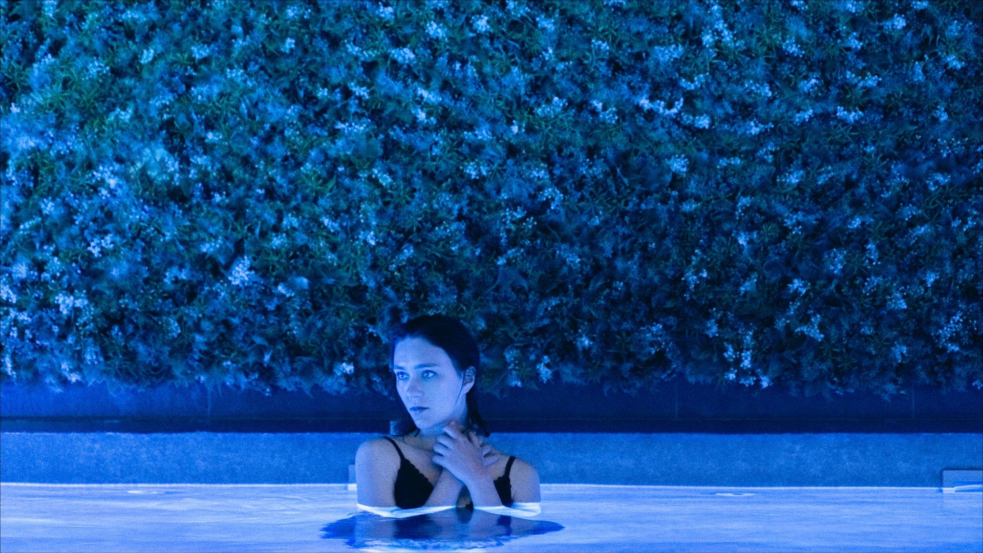 Woman in a pool at night, hands on her neck, looking up with a worried expression. 