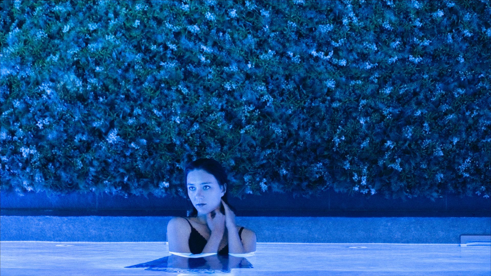 Woman in a pool, lit with blue light, hands near her face, looking at the camera. Green foliage in background.