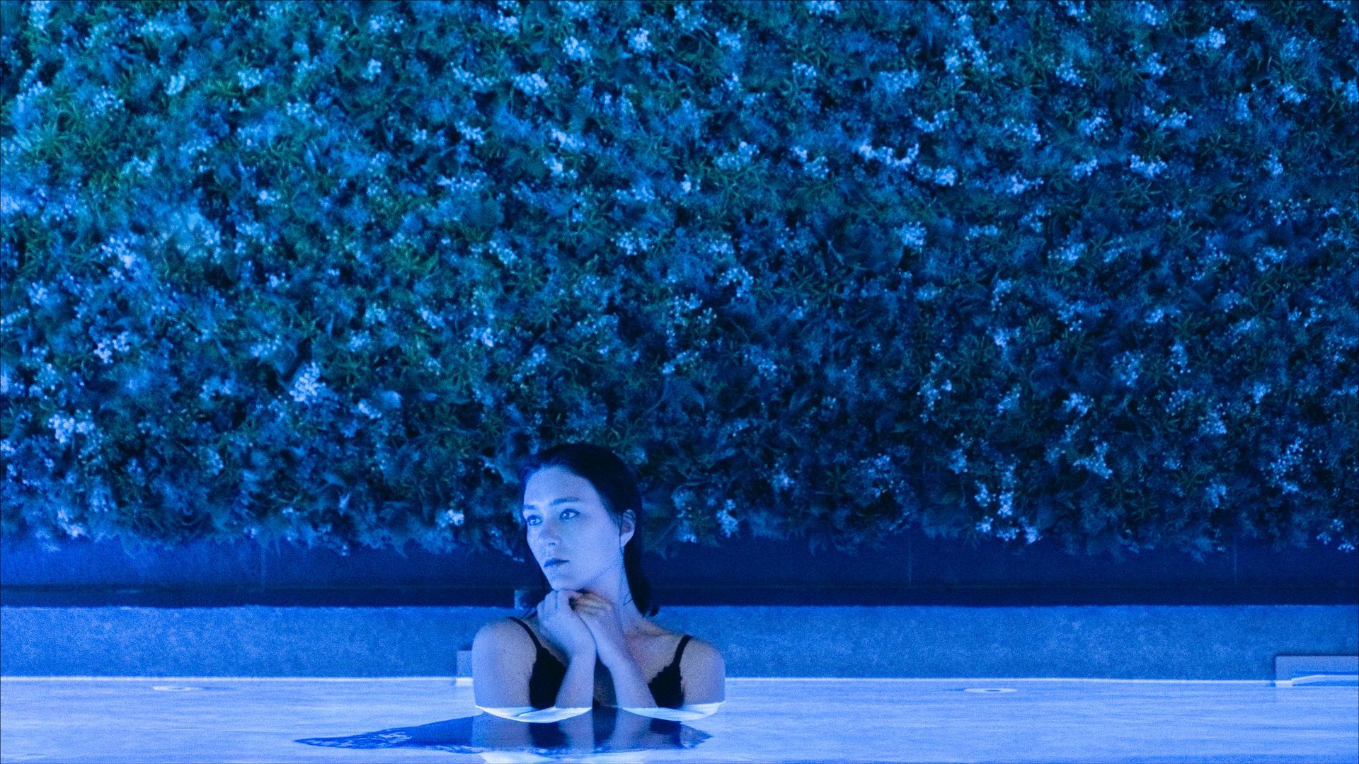 Woman in a pool, hands bound, looking up with a surprised expression, in front of a blue-lit hedge.