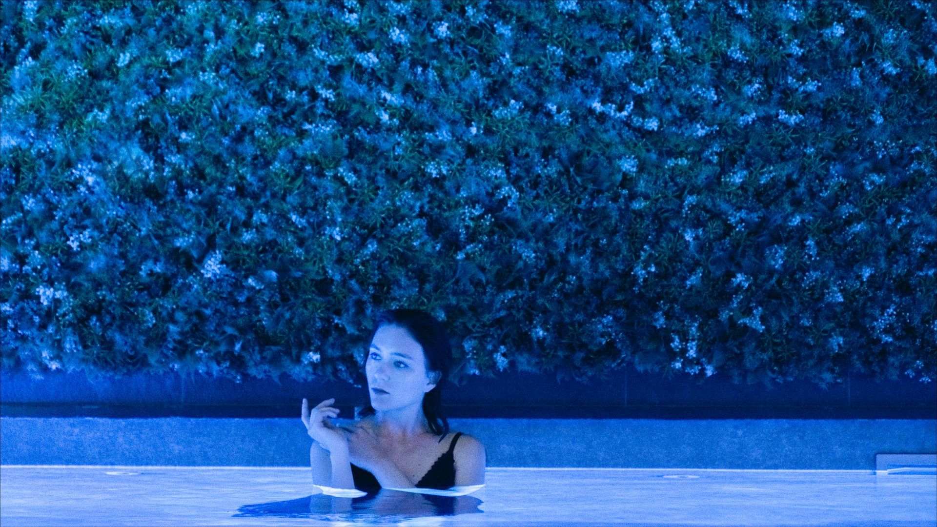 Woman in a pool at night, looking up. Illuminated by blue light, a wall of green foliage behind.