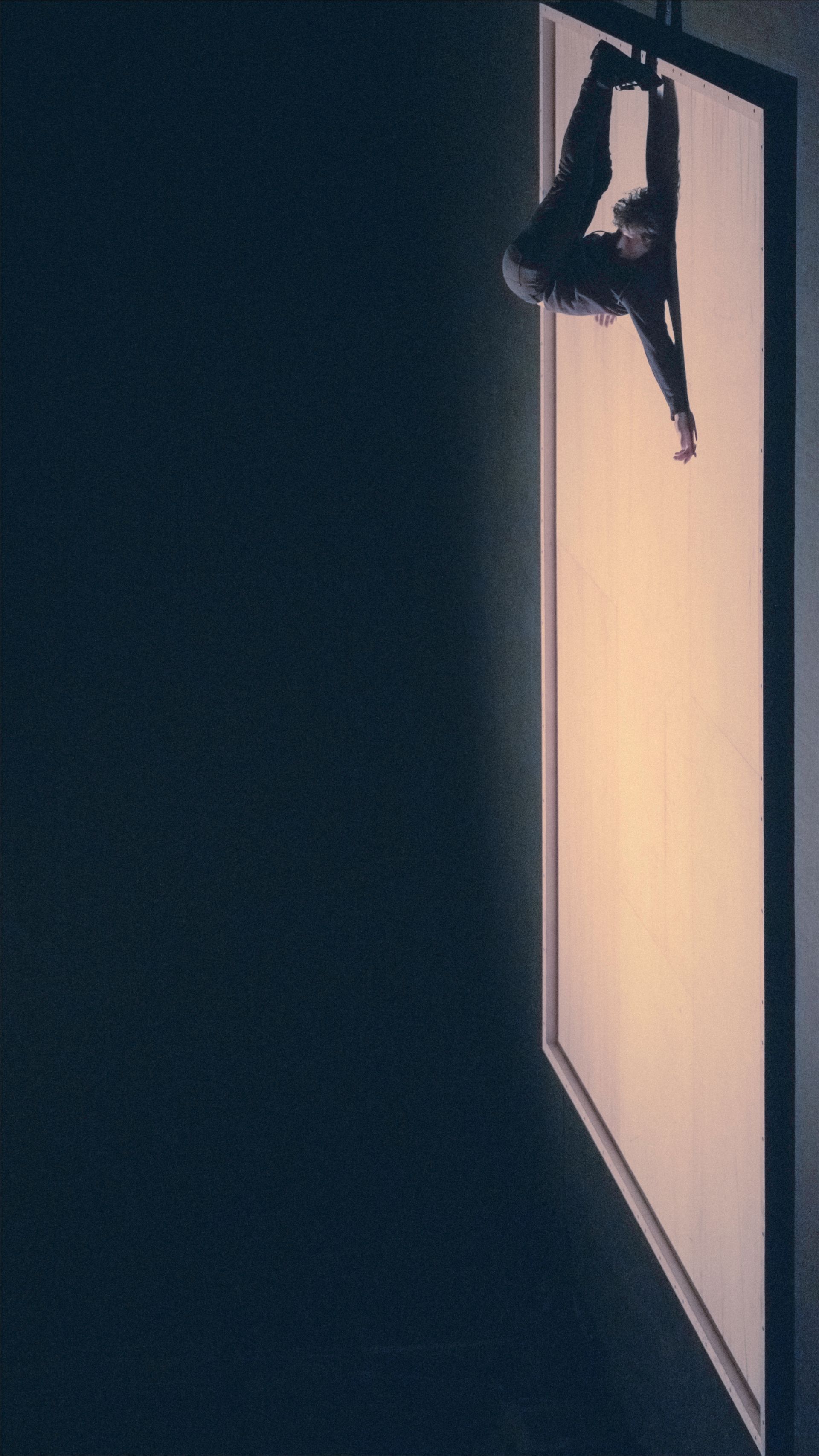 Person hanging from a large, illuminated screen in a dark space, arm outstretched.