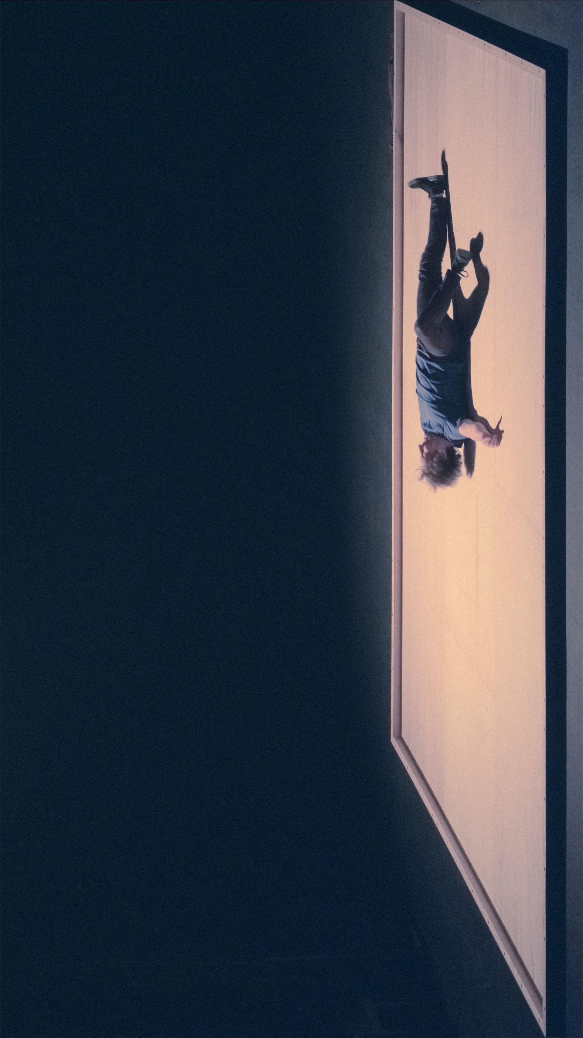 Person hanging upside down on a bright wall, dark background.