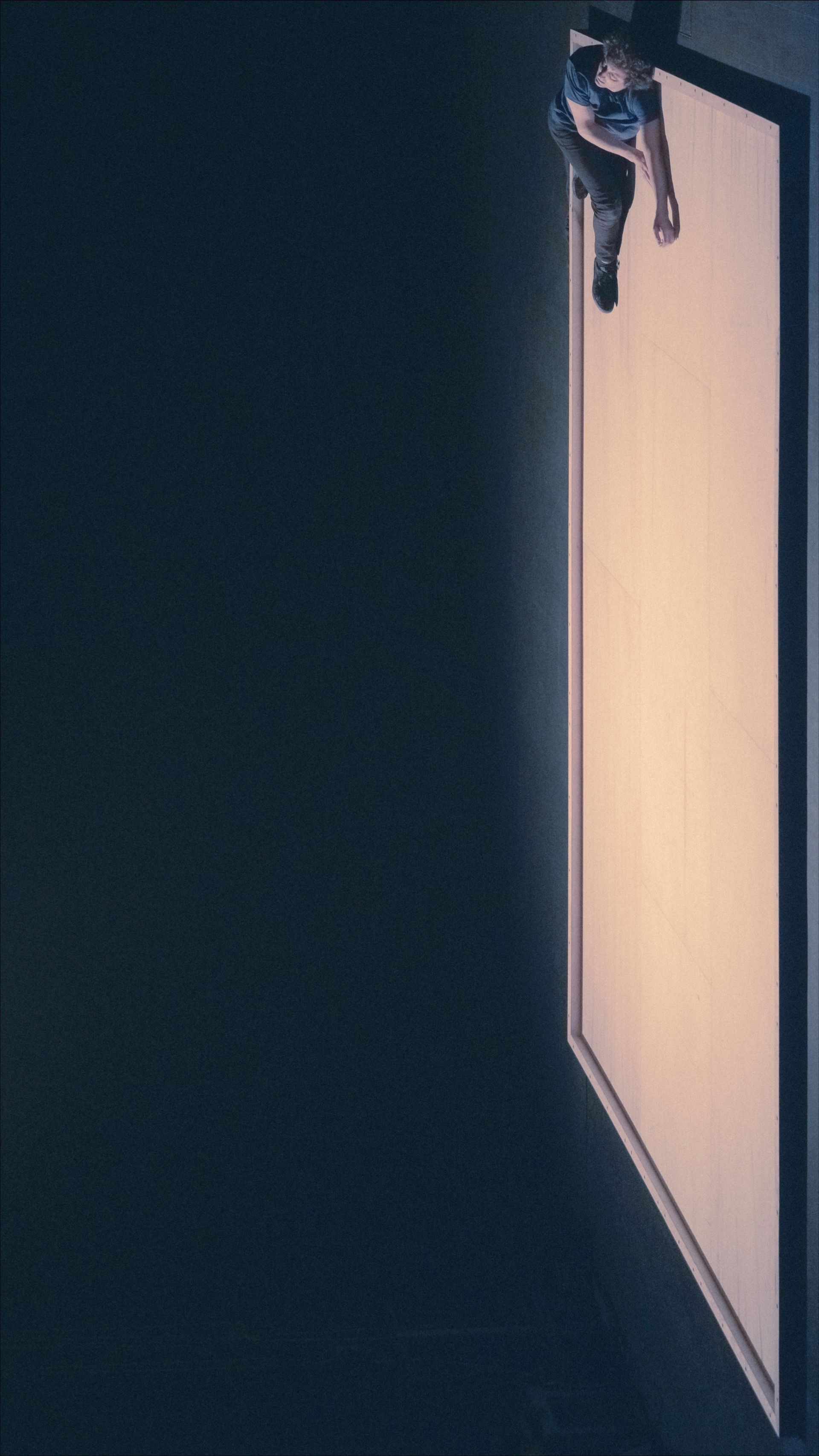 Person hanging off the edge of a bright, rectangular structure against a dark background.