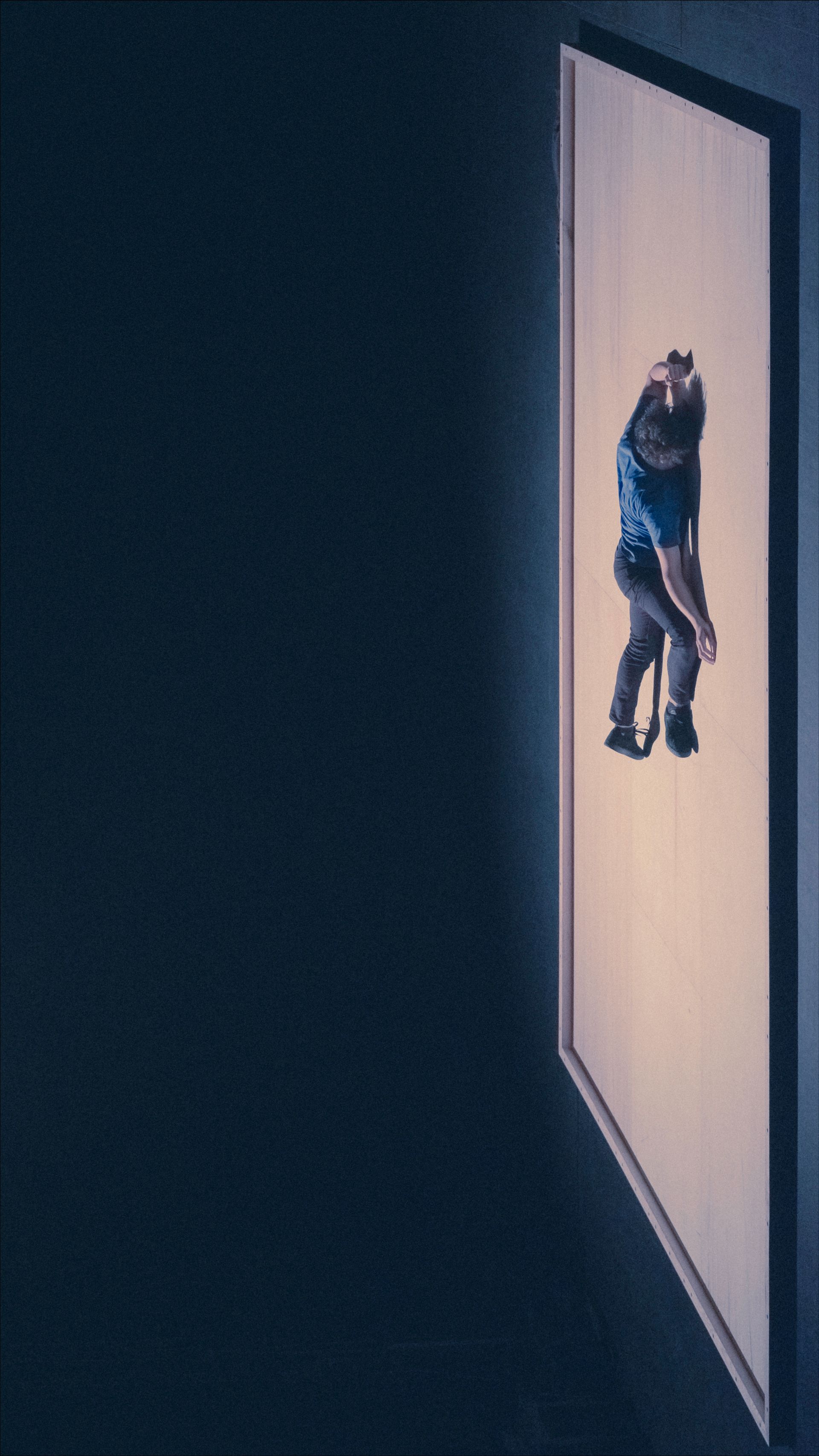 Person clinging to the side of a tall, light-colored rectangular wall in a dark setting.