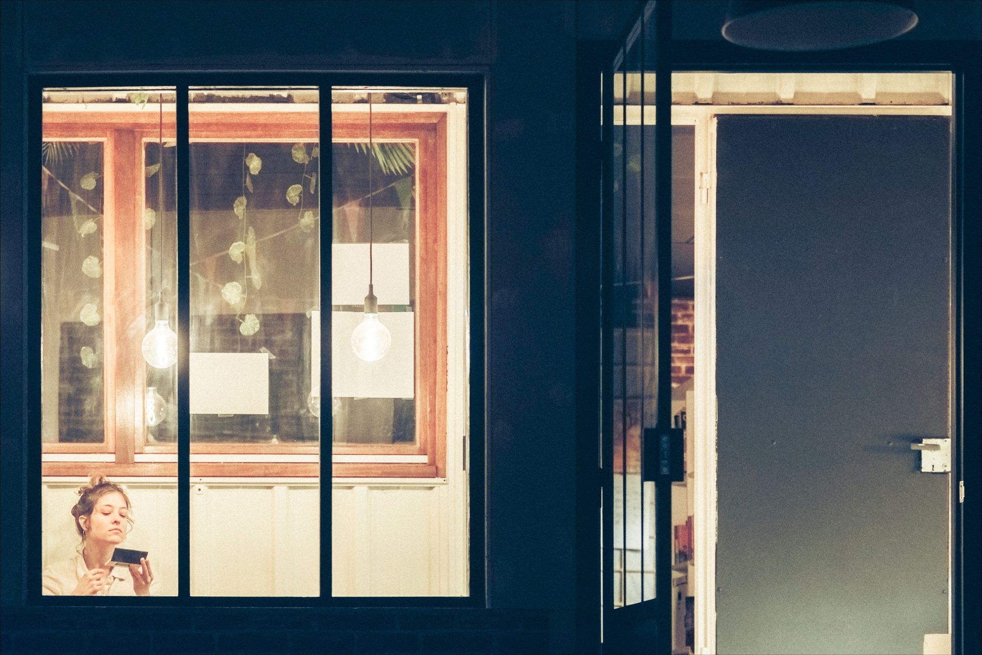 Woman looking in a mirror by a window at night. Interior door open on the right.