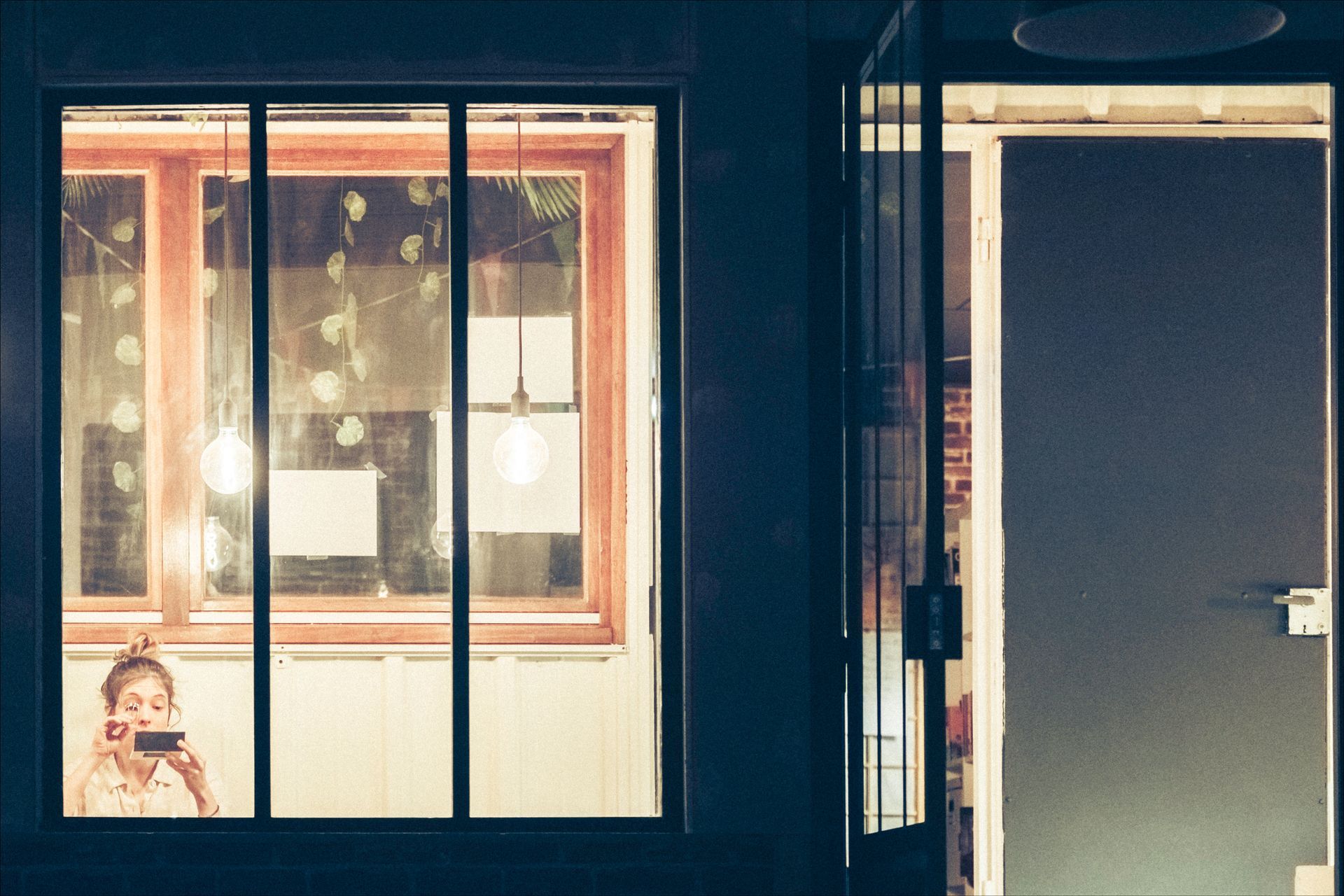 Woman taking photo with phone, seen through window at night; warm interior light.