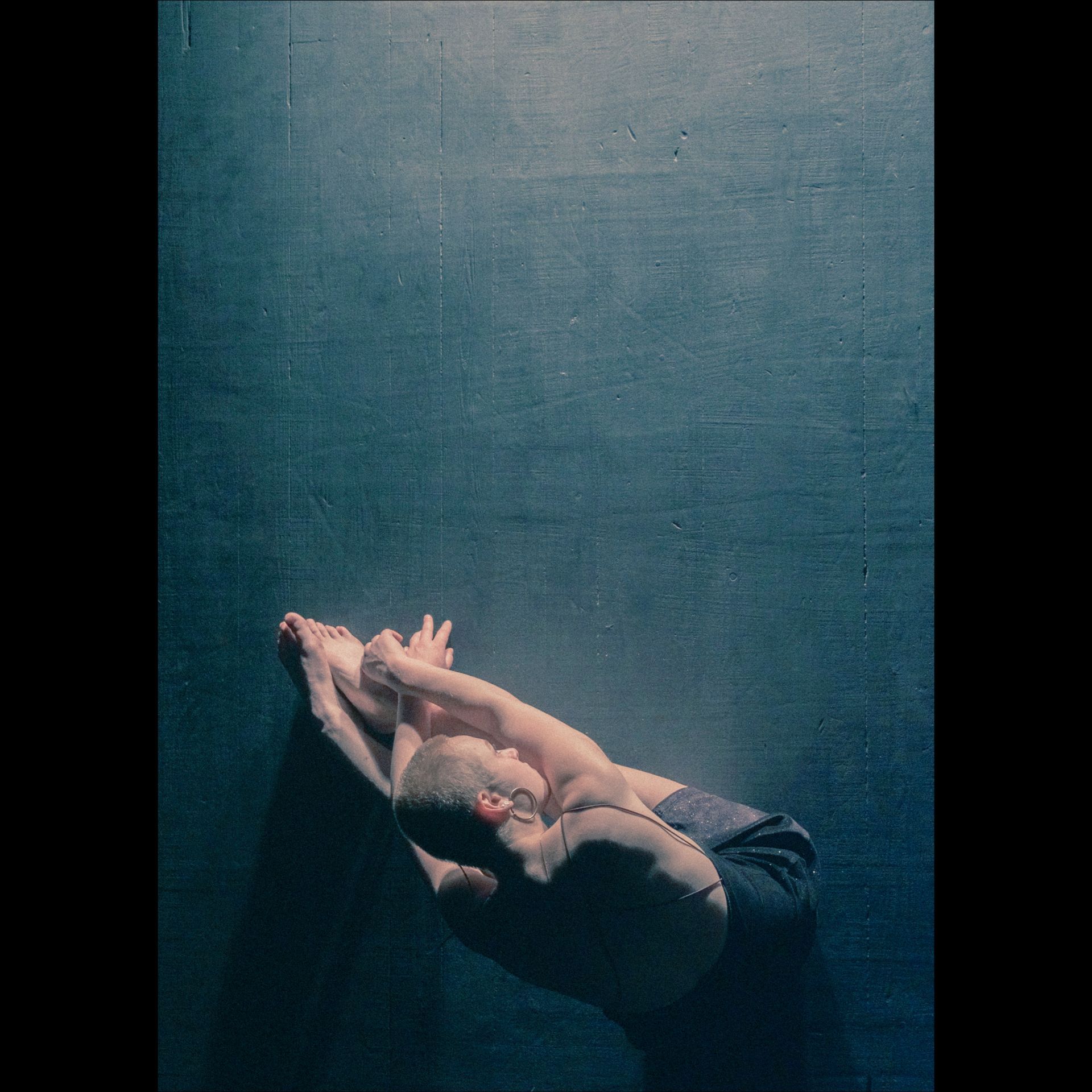 Person in a seated forward bend, torso curved over legs, hands on feet, against a dark blue wall.