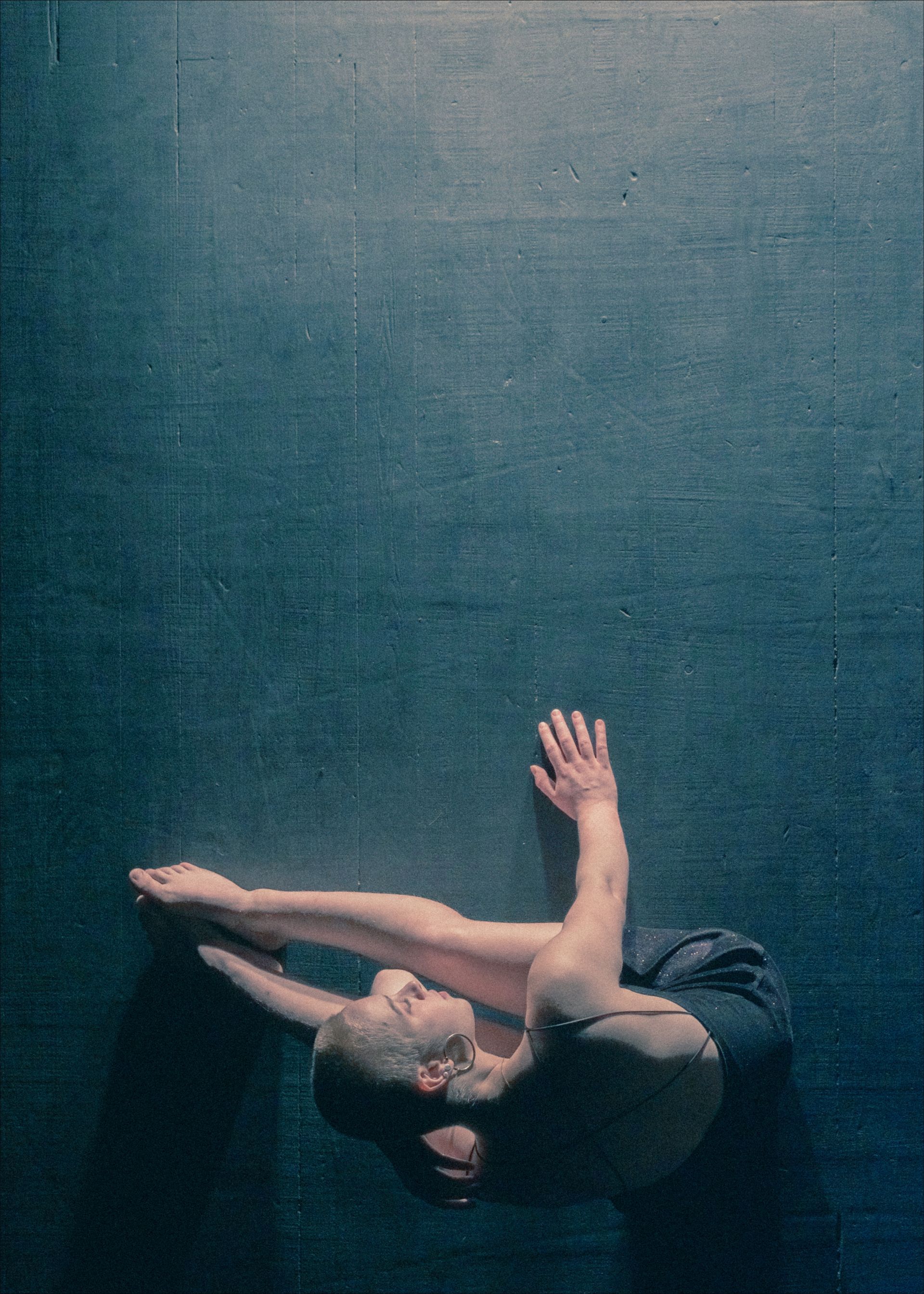 Dancer on floor, curled in a pose. Arms extended, head turned. Dark setting, blue tone.