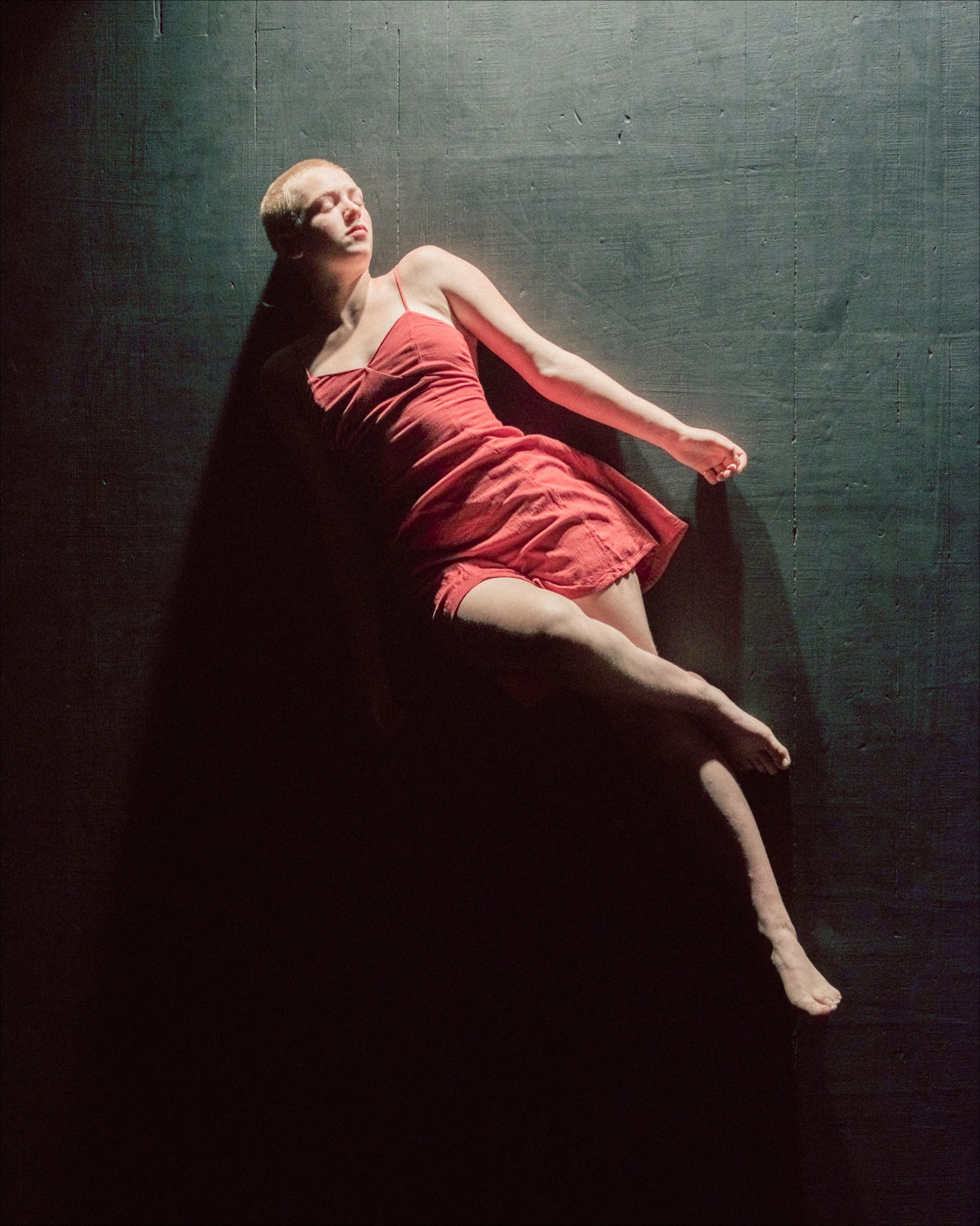 Woman in red dress lies on a dark surface, arms extended, lit by a spotlight.