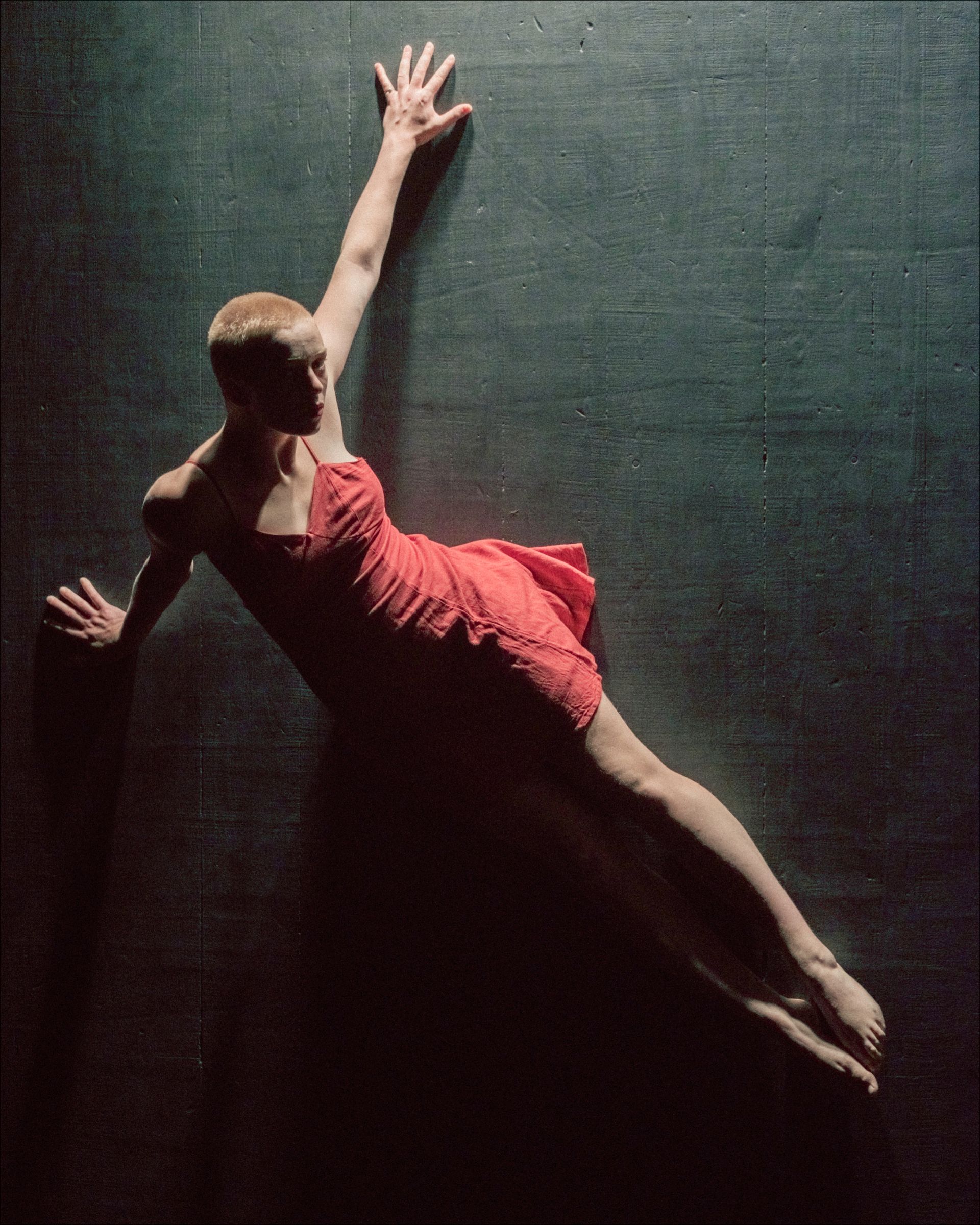 Dancer in a red dress, leaning against a dark wall with one arm extended upwards.