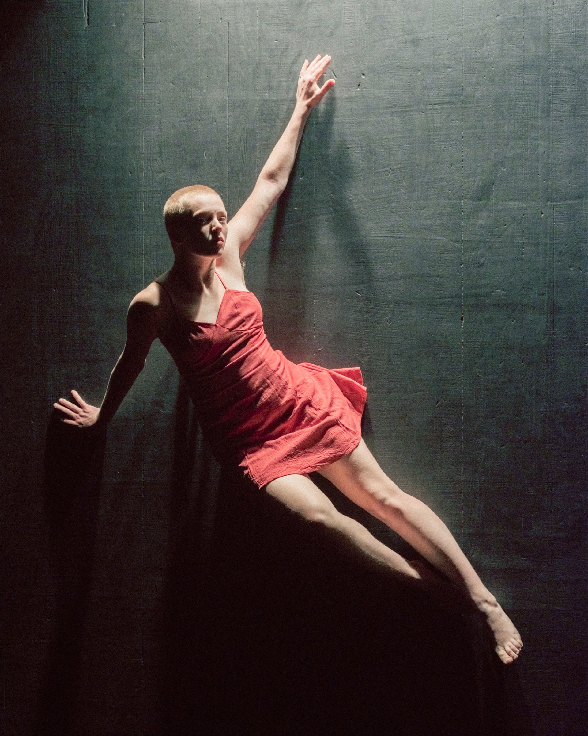 A person with a shaved head in a red dress performs a dance pose against a dark wall.