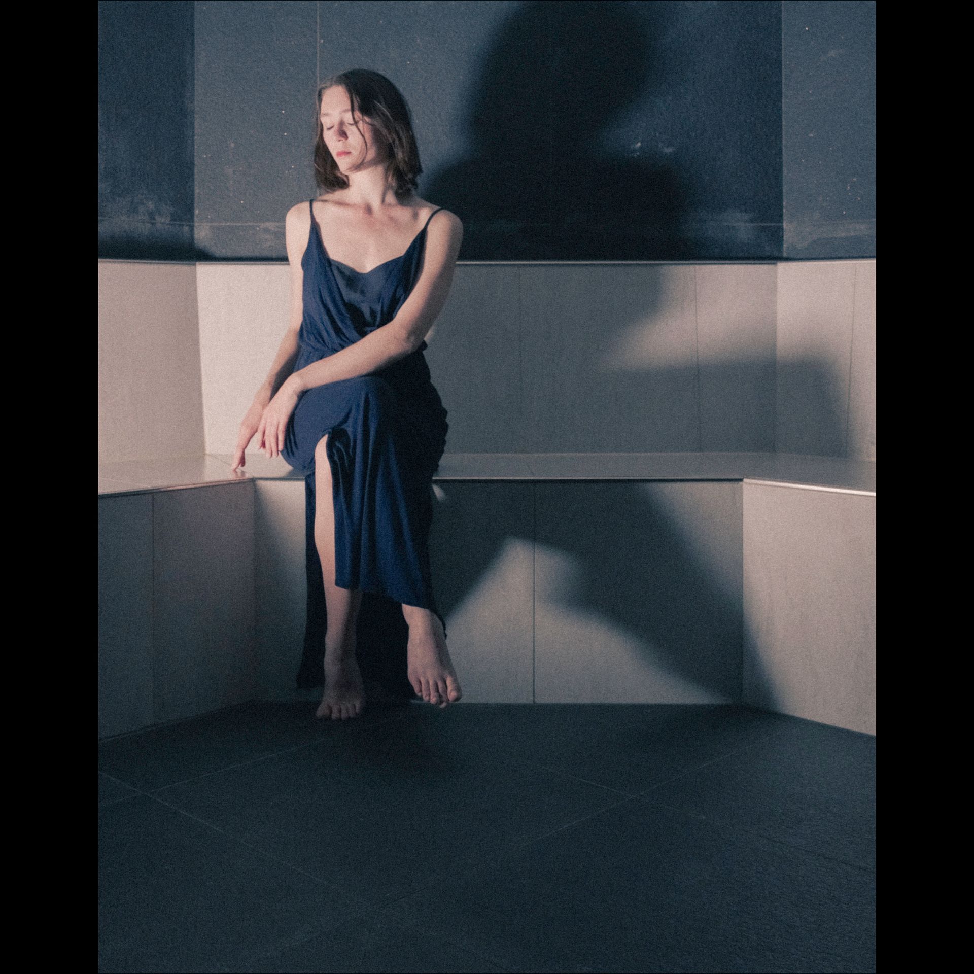 Woman in blue dress sits on a bench, looking down. Dark room, shadow on wall.