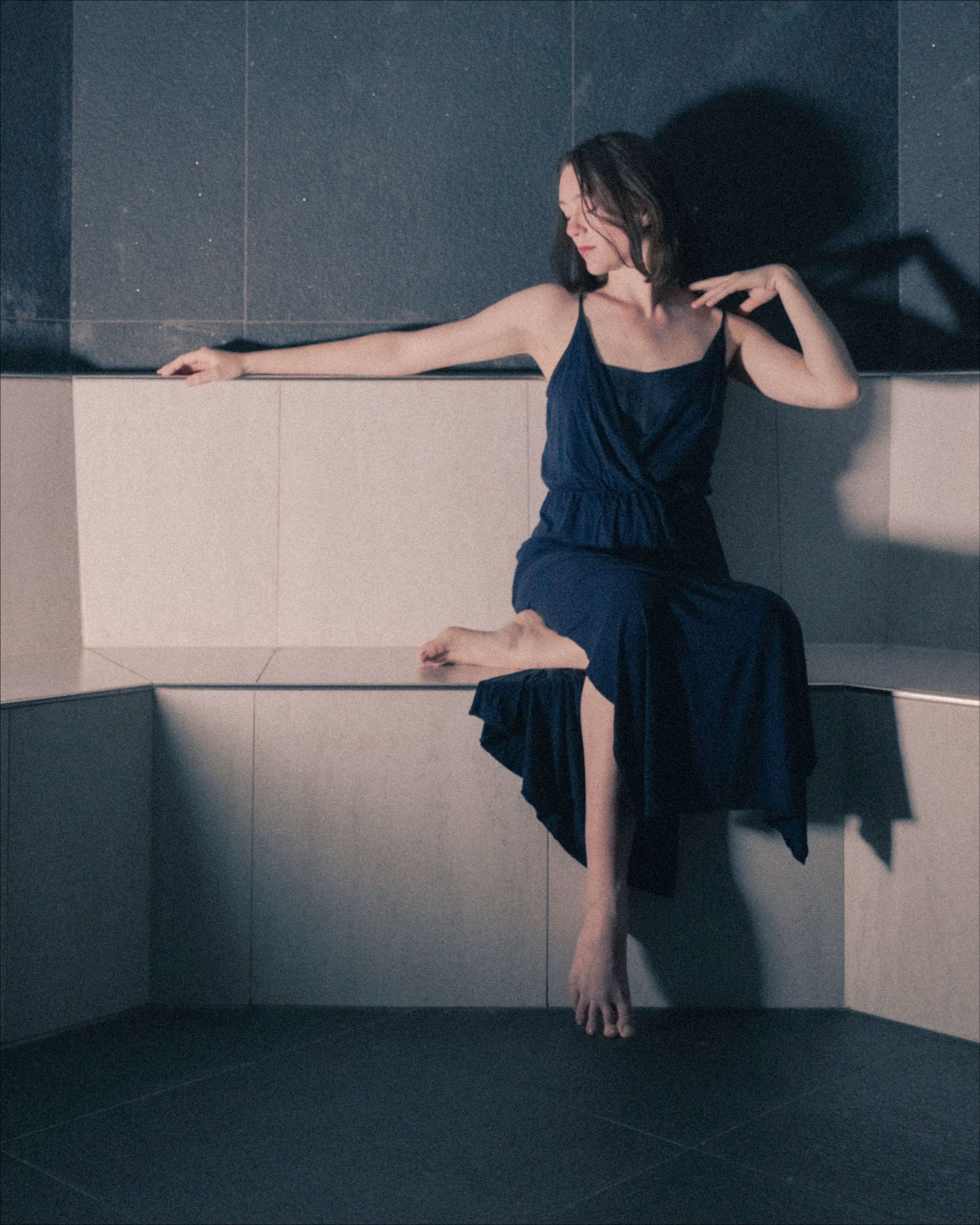 Woman in blue dress sits on a tiled bench, barefoot, in a modern, gray-toned space.