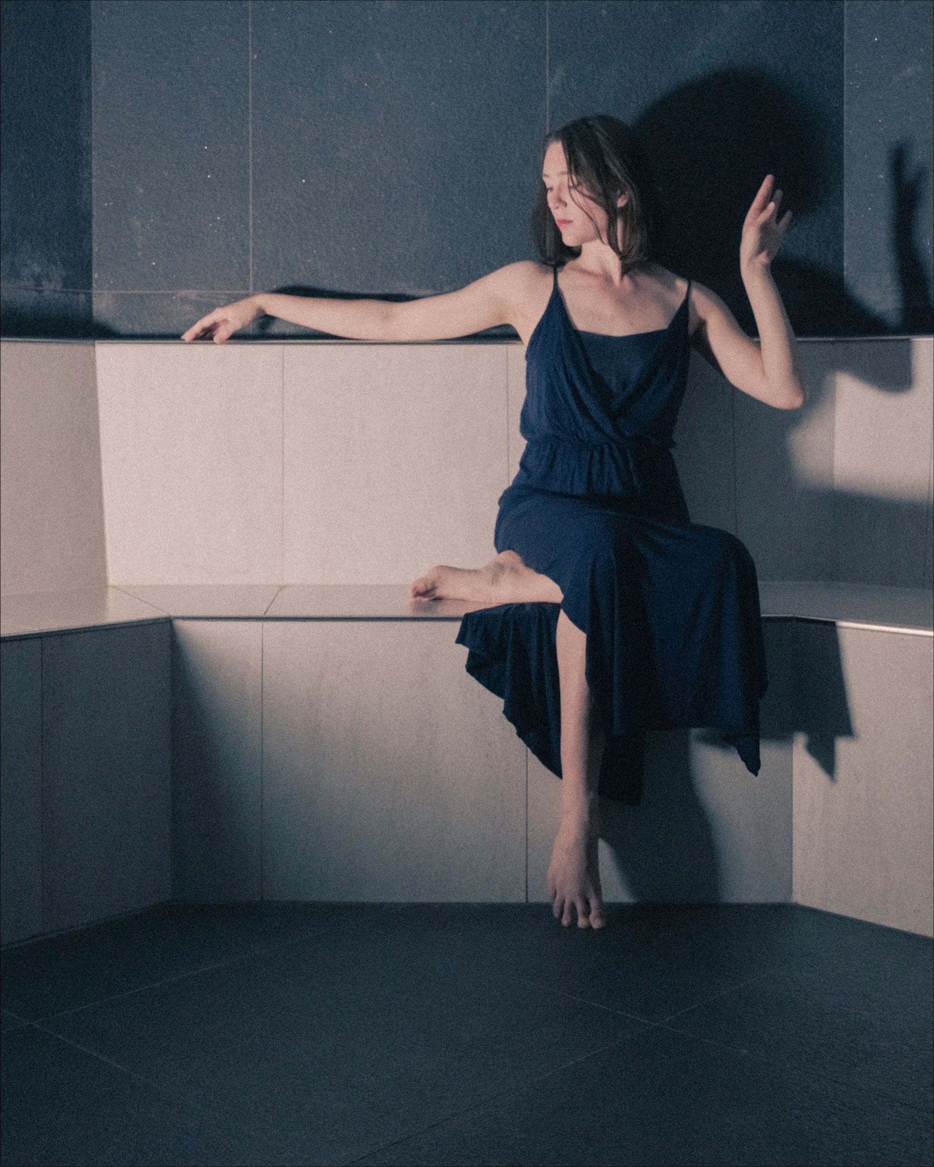 Woman in blue dress sits on a tiled ledge, one arm extended, other raised. Dark setting.