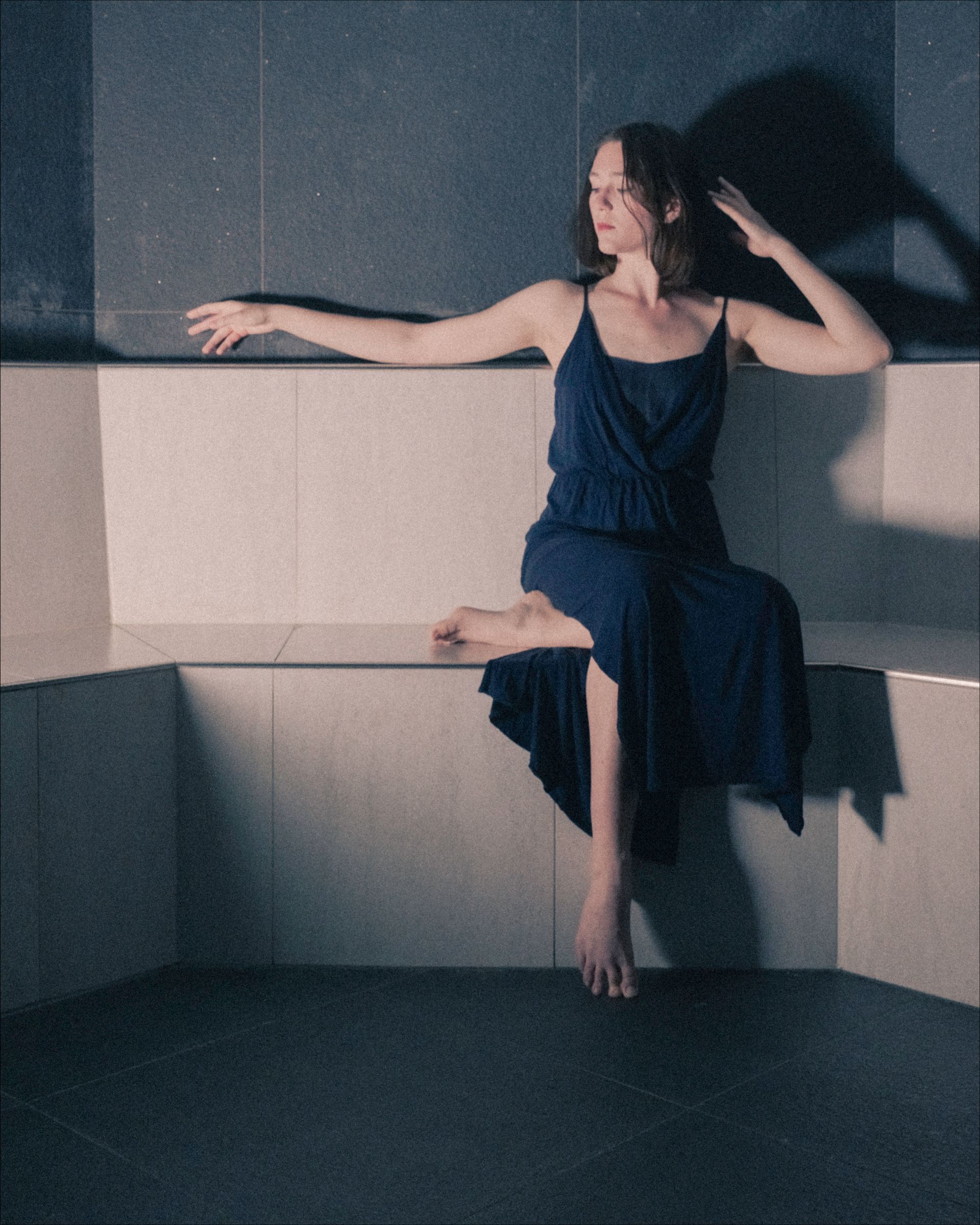 Woman in blue dress poses on a raised, white bench, one arm extended. Dimly lit interior.