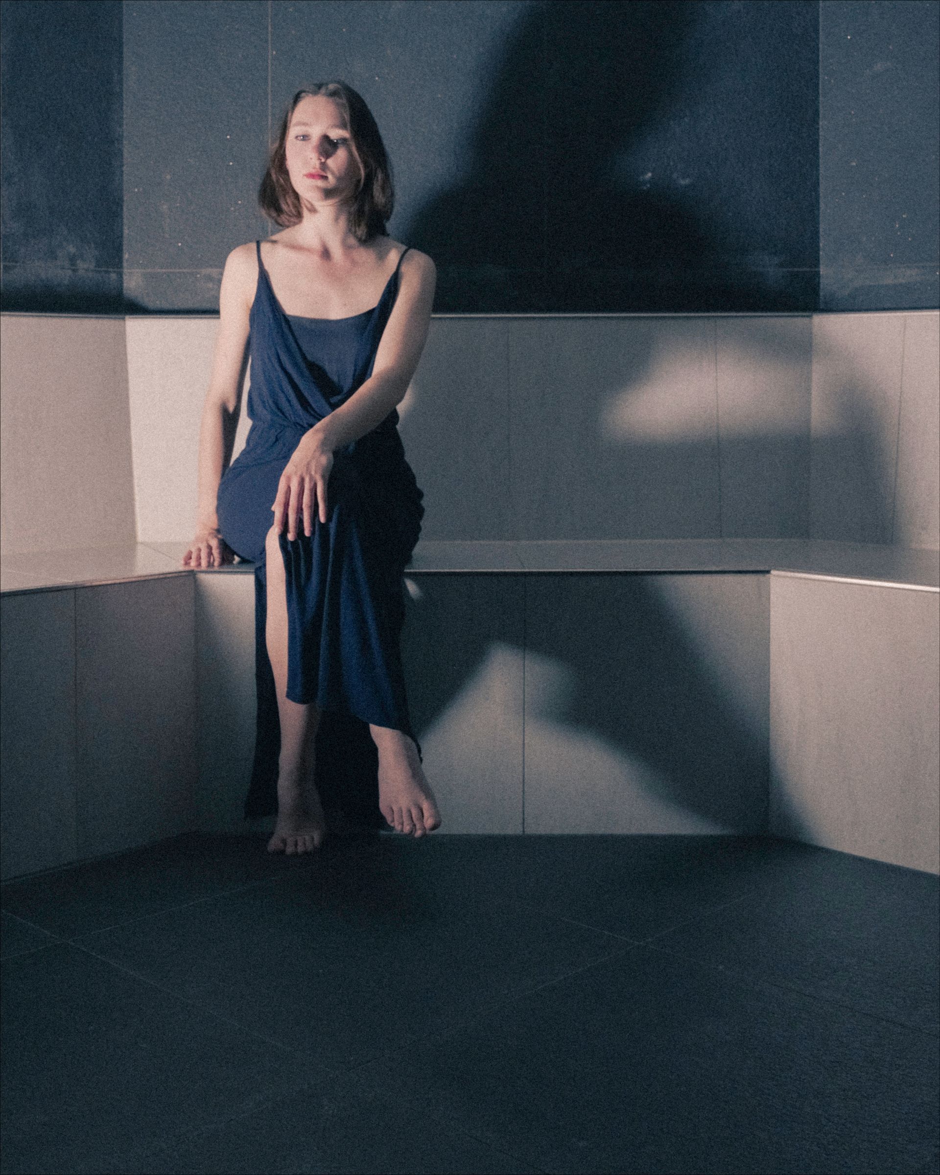 Woman in blue dress sits on a tiled bench, looking off-camera. Shadow behind.