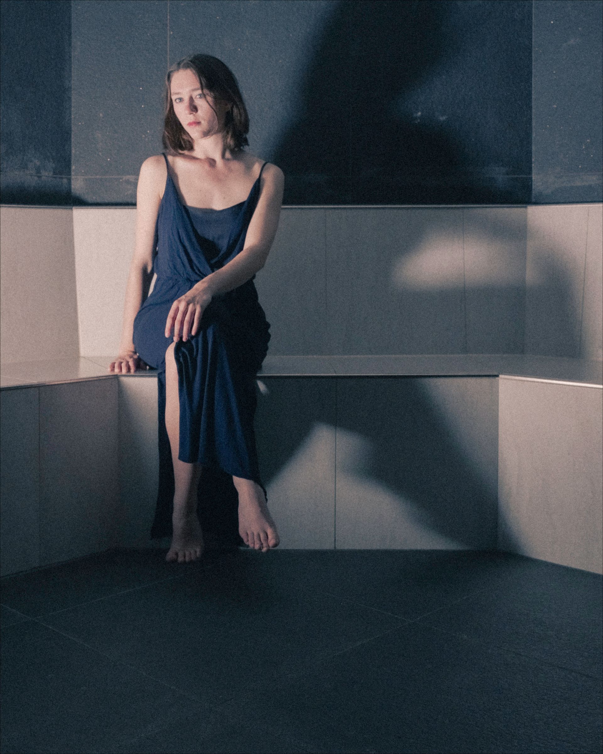 Woman in blue dress sits on a concrete ledge, looking downward. Moody lighting and shadow.
