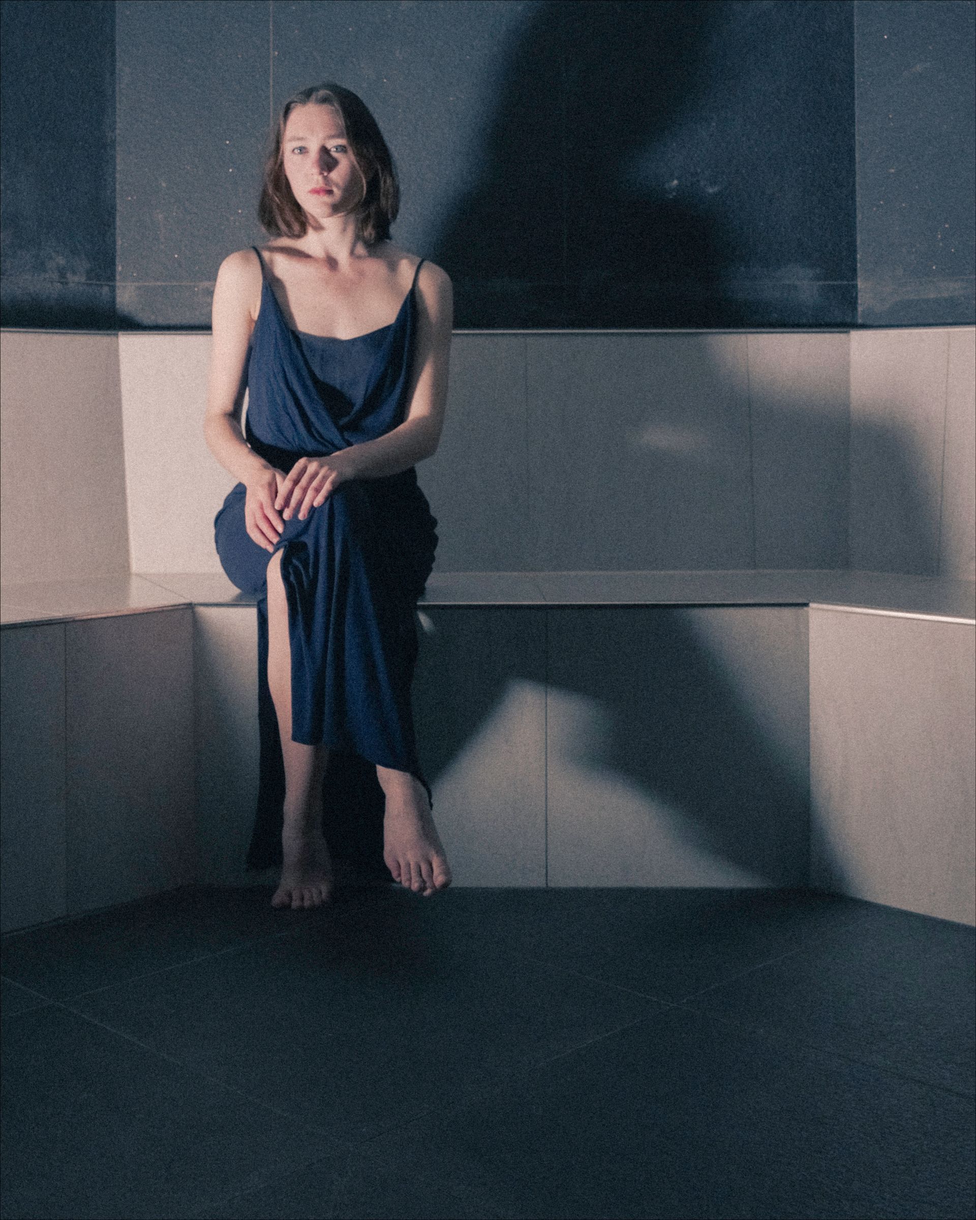 Woman sitting in a tiled alcove, wearing a blue dress. A shadow stretches across the wall.