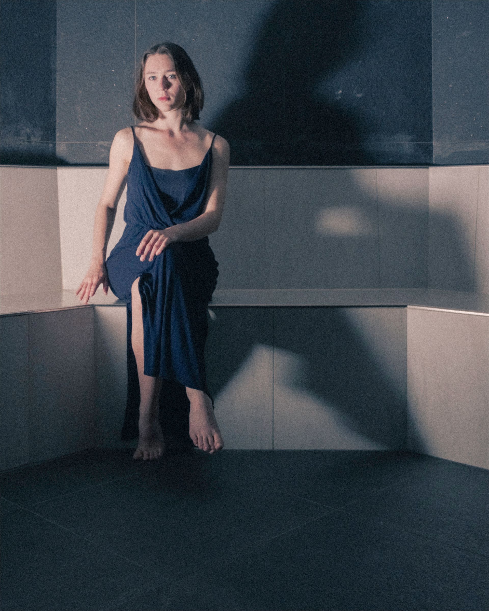 Woman in blue dress sits on a gray ledge, looking at the viewer. Soft shadows.