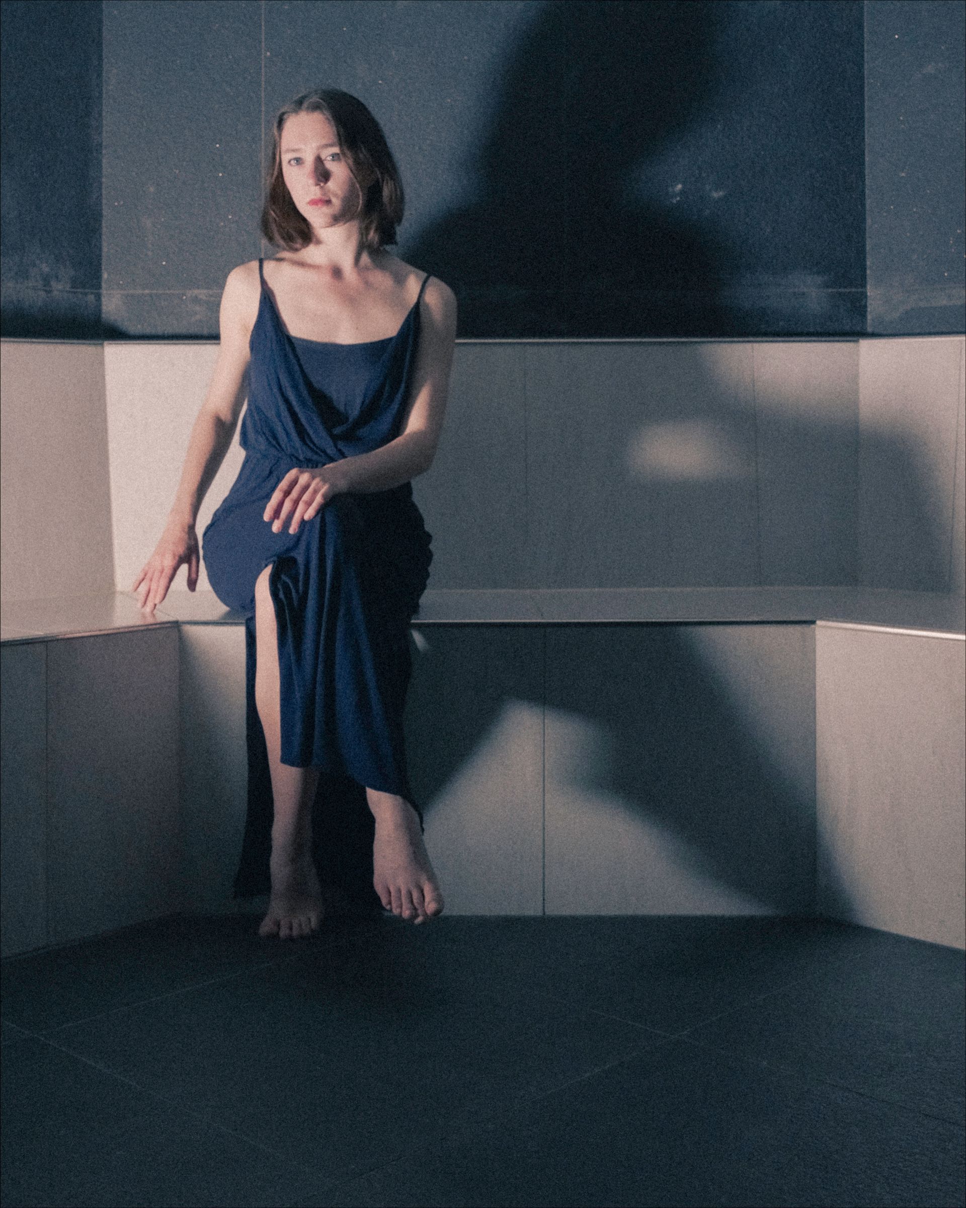 Woman in blue dress sits on ledge in a shadowy, minimalist room.