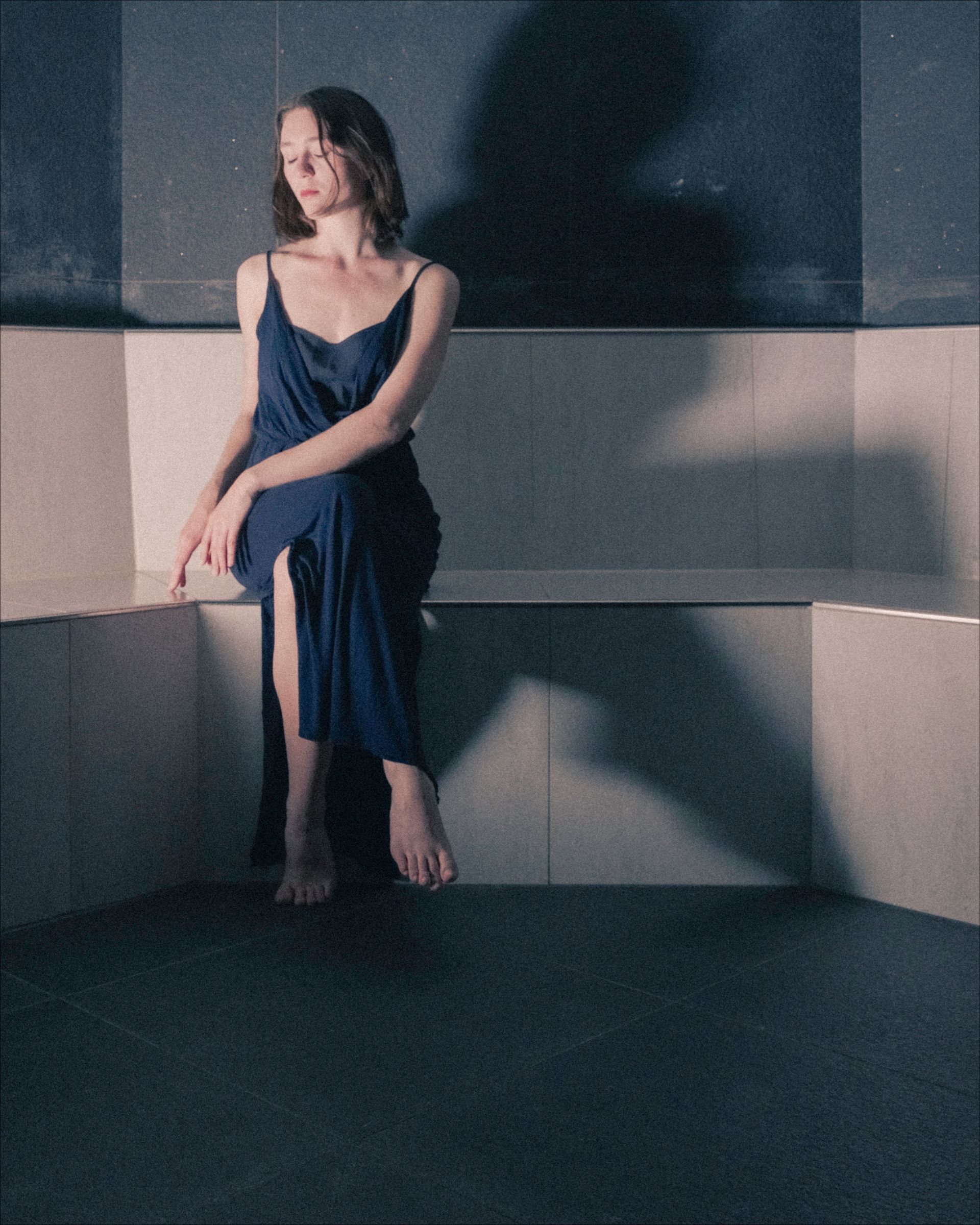 Woman in blue dress sits on a bench, shadowed walls, barefoot.