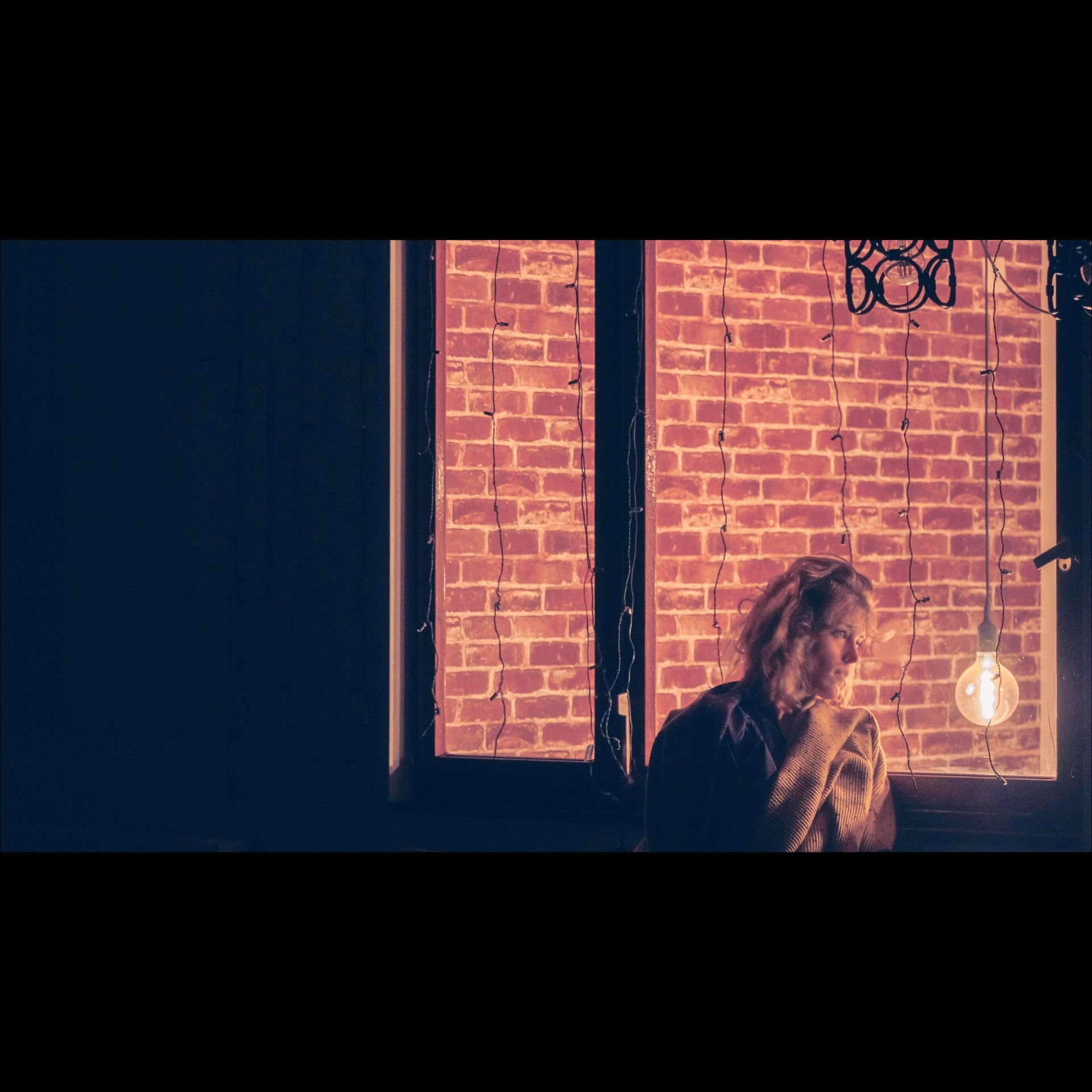 Person by a window with exposed brick, illuminated by a hanging light. Dark space on left.