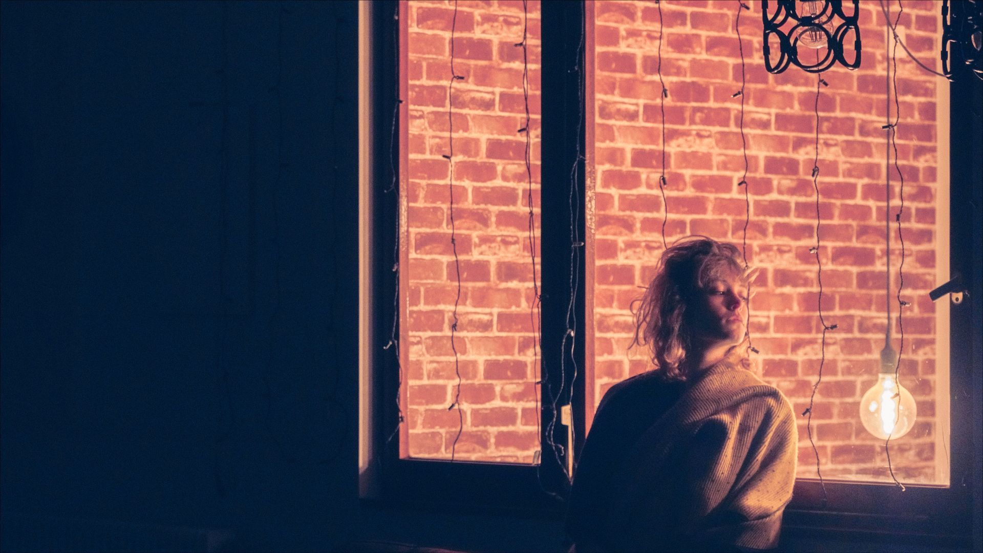 Person in a robe stands near a window with a brick wall backdrop. A hanging light bulb illuminates the scene.