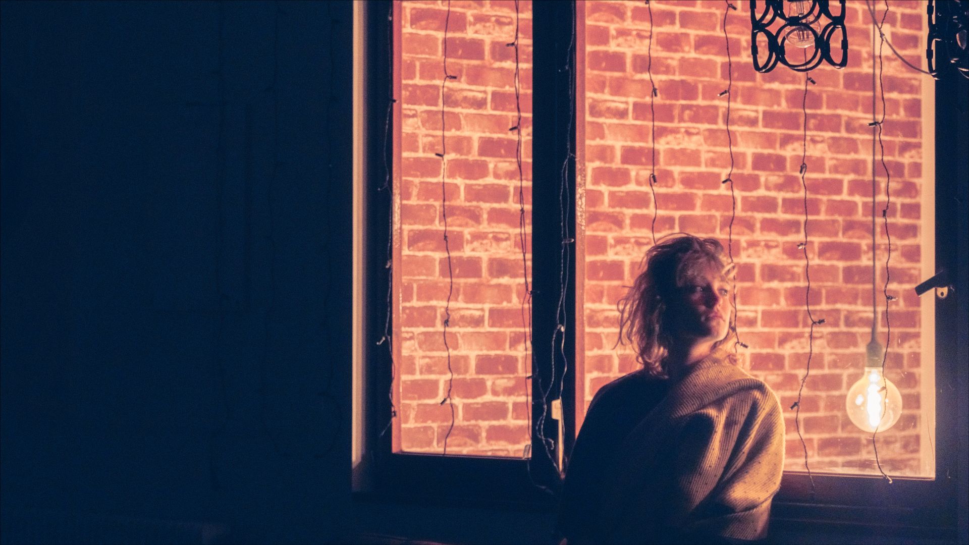 Person looking out a window at a brick wall lit by a warm glow. A single light bulb hangs nearby.