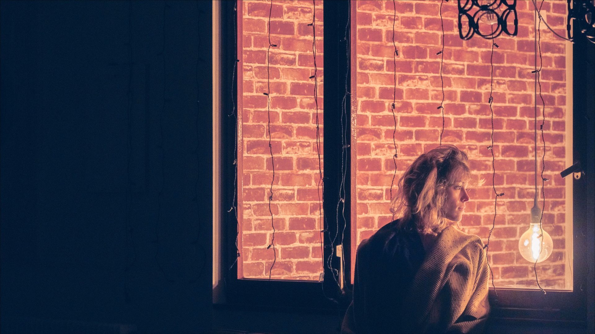 Person looking out a window with brick wall, illuminated by a hanging light bulb, in a dimly lit room.