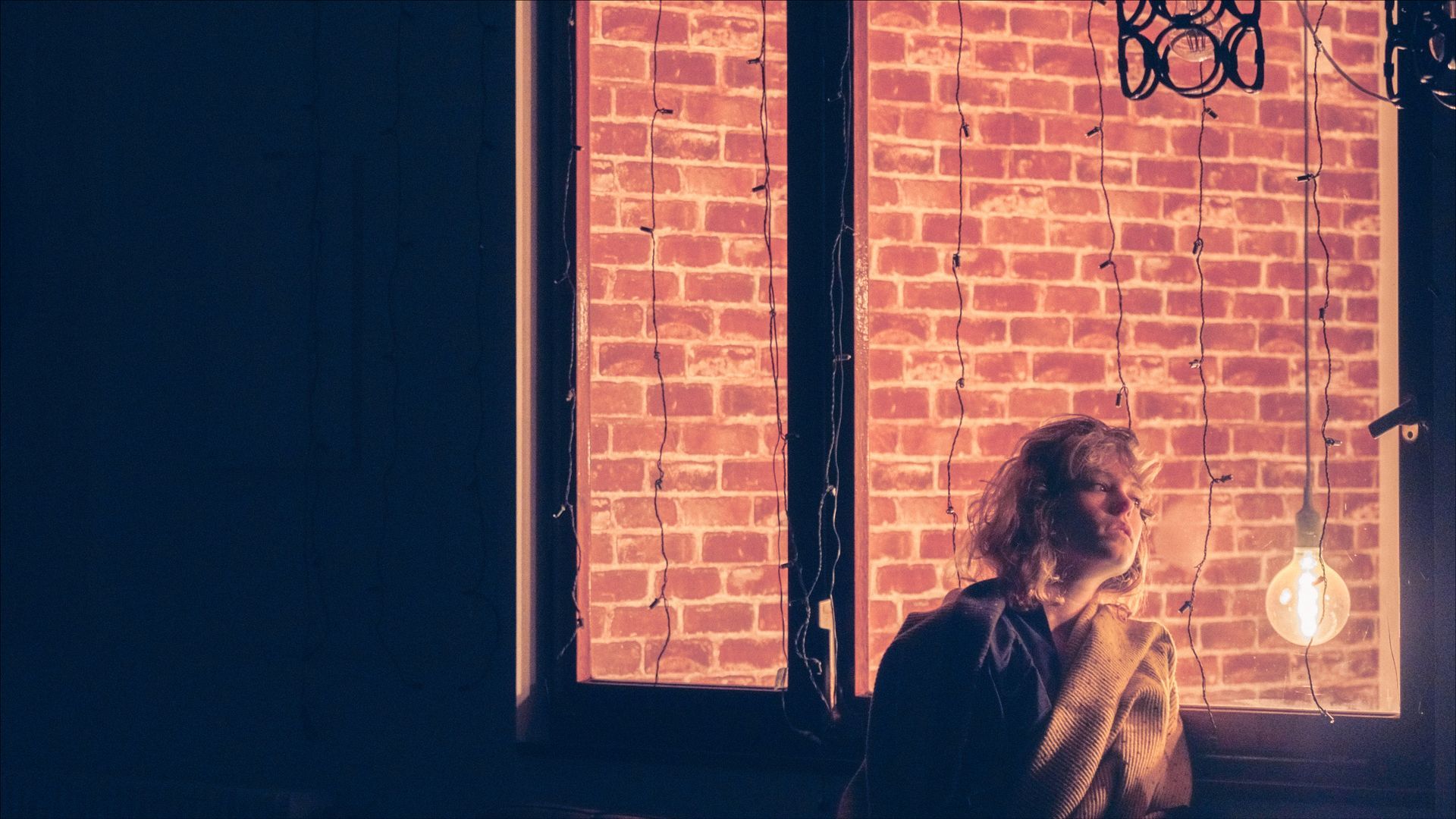 Person looking out a window with brick wall, illuminated by a hanging light bulb. Orange-toned lighting.