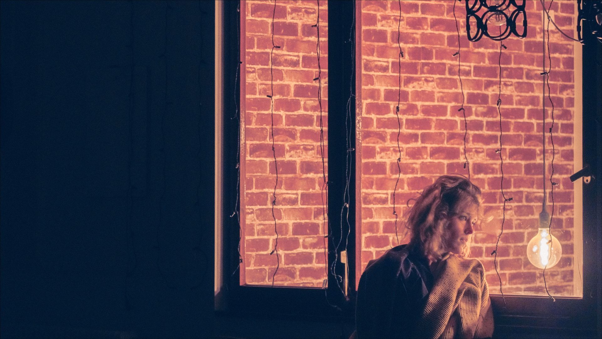 Person by a brick wall window, lit by a hanging bulb. Dark interior, natural light.