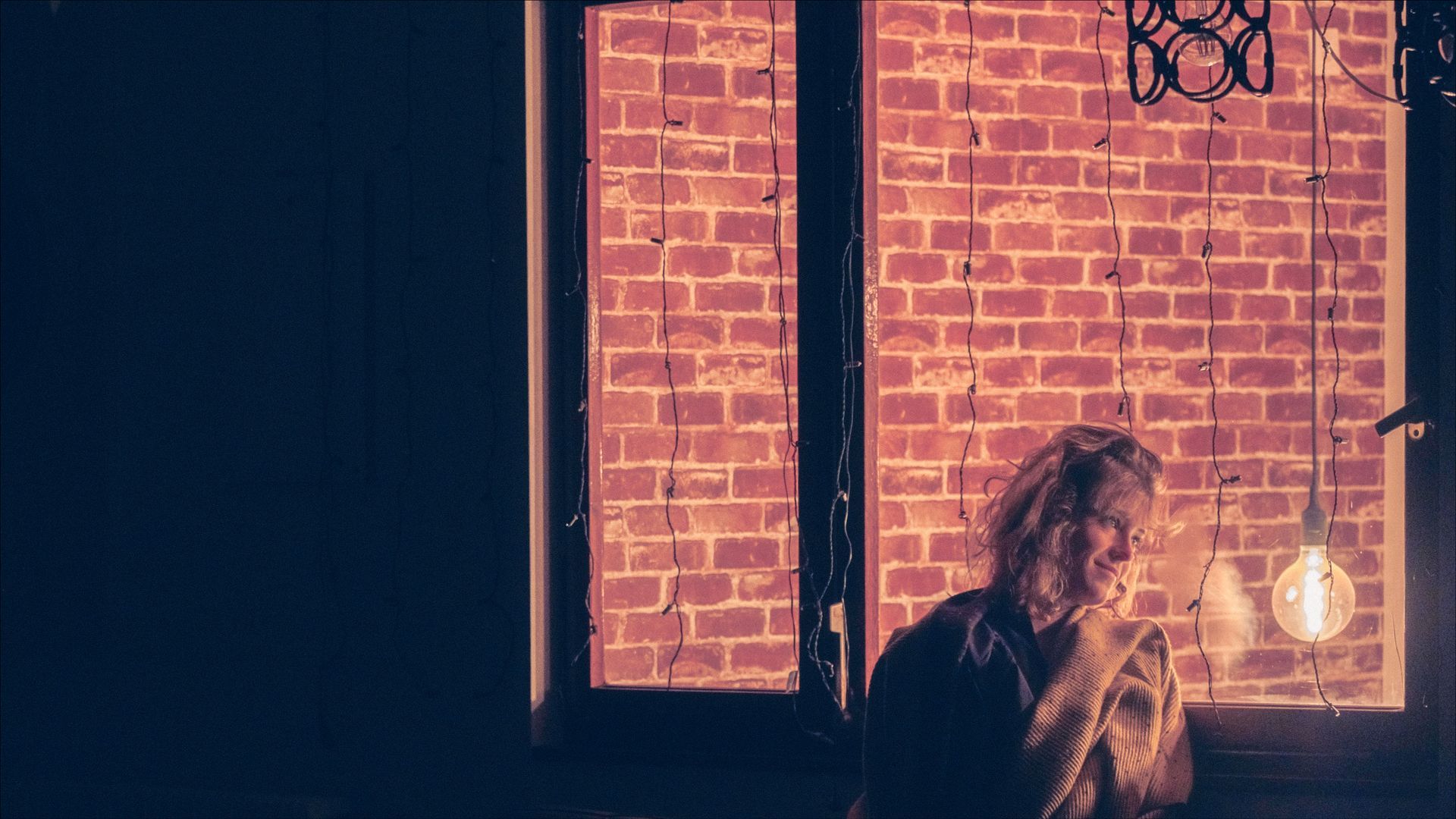 Person near a window with exposed brick wall, lit by a hanging bulb; dimly lit.