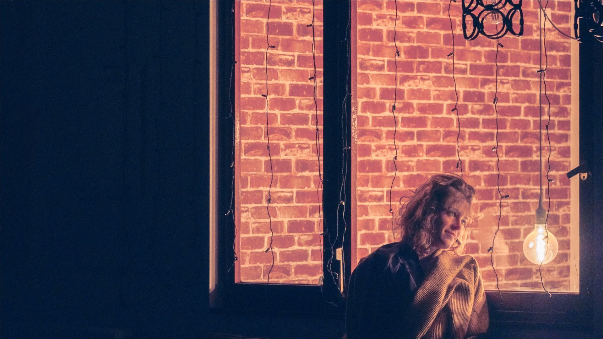 Person near window, lit by a hanging bulb. Brick wall in background, warm tones.