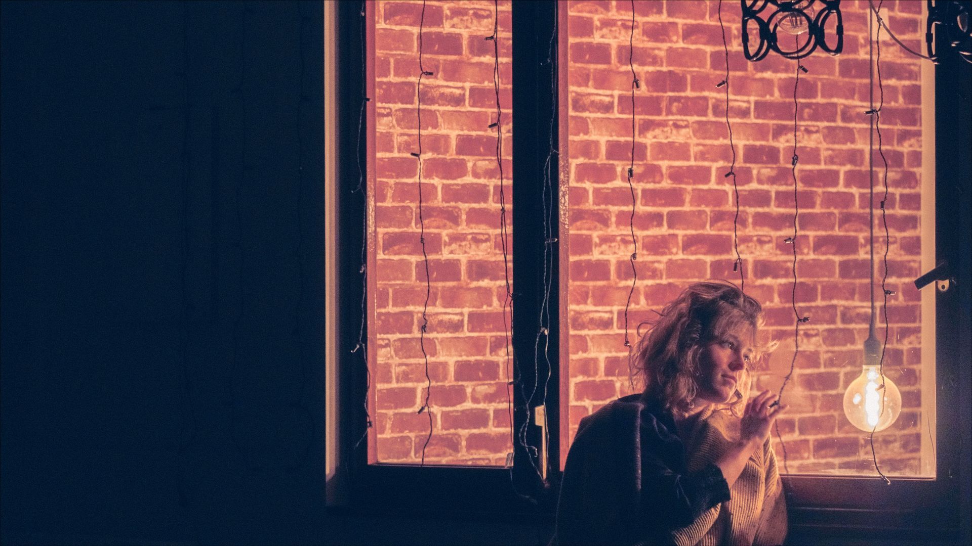 Person near a brick wall window with a hanging lightbulb. Dimly lit room, holding a cigarette.