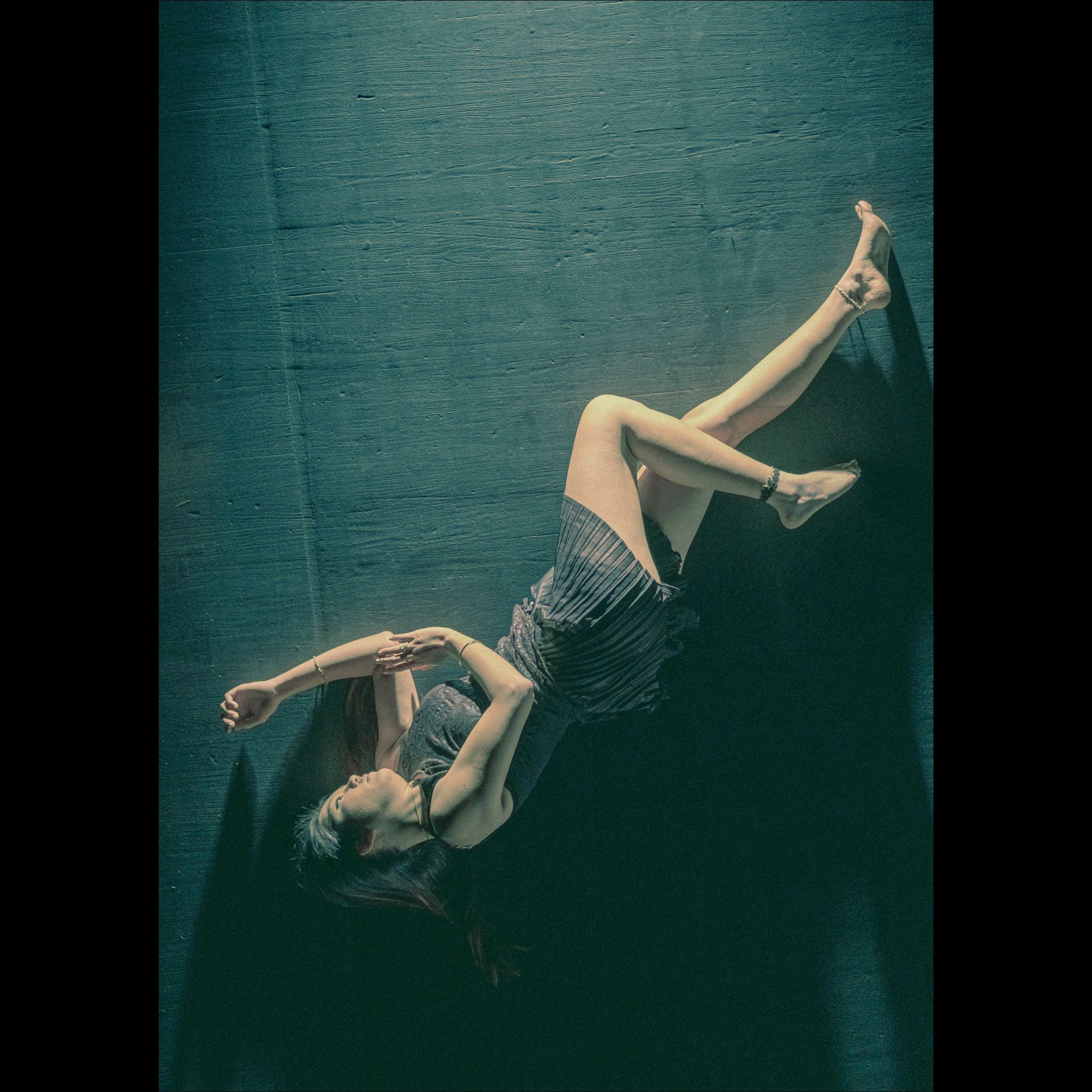 Woman in a dark dress, lying diagonally on a textured teal surface with arms outstretched.