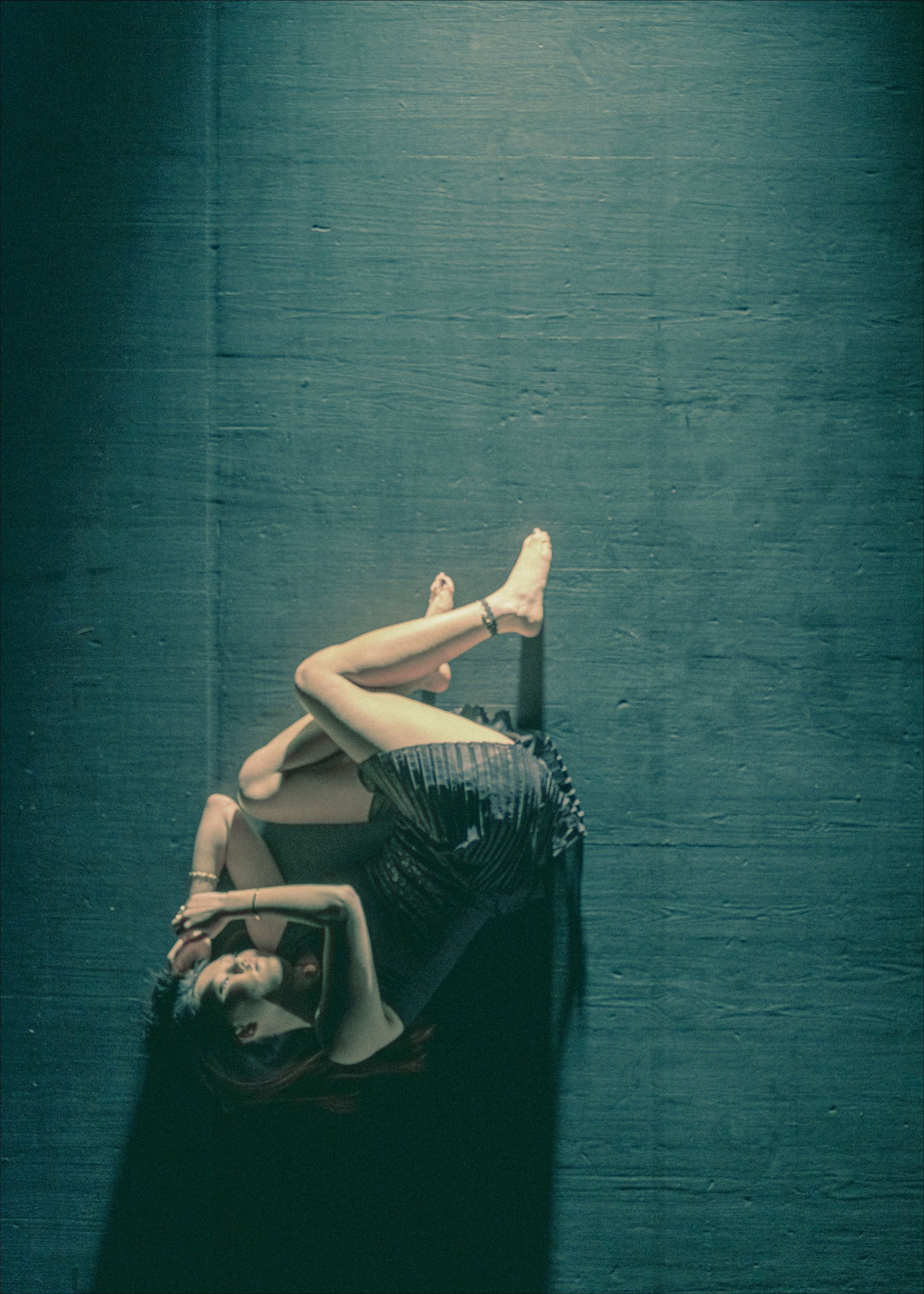 A person lies on a dark blue floor, legs bent, arms overhead. Their skin is light, and they wear a dark, short dress.