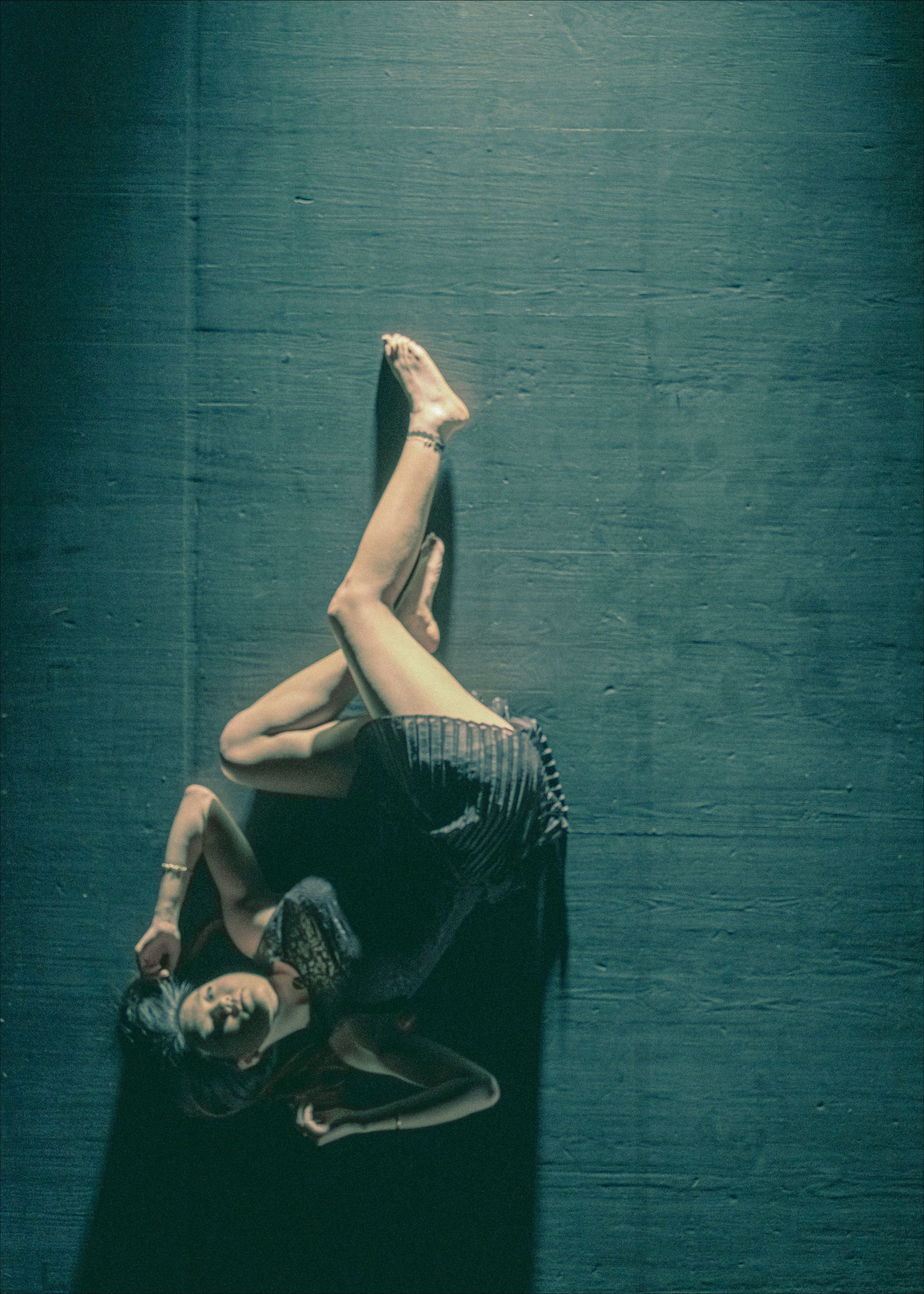 Dancer on floor in dark room. Legs crossed, body arched, arms bent; dark clothing, focused expression.